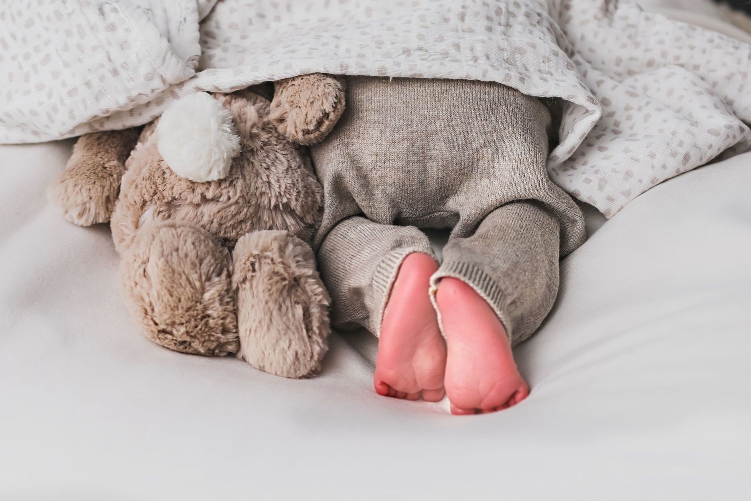 A plush teddy bear and pink bunny slippers lay on white bedding.