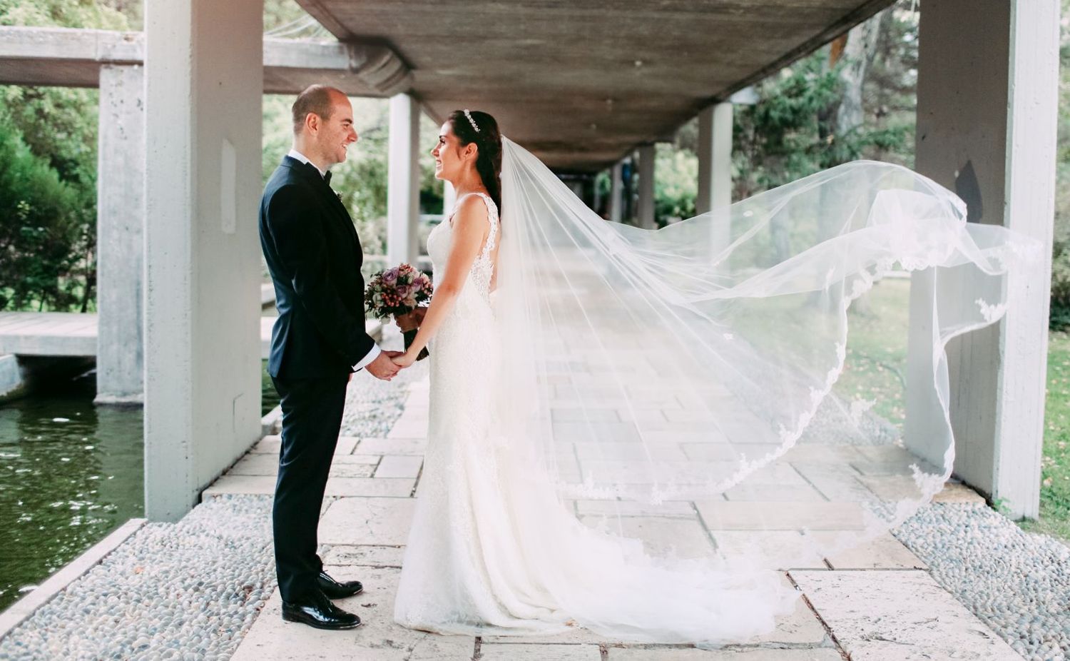 A wedding couple stands under a modern architectural overhang with a flowing veil caught in the breeze.