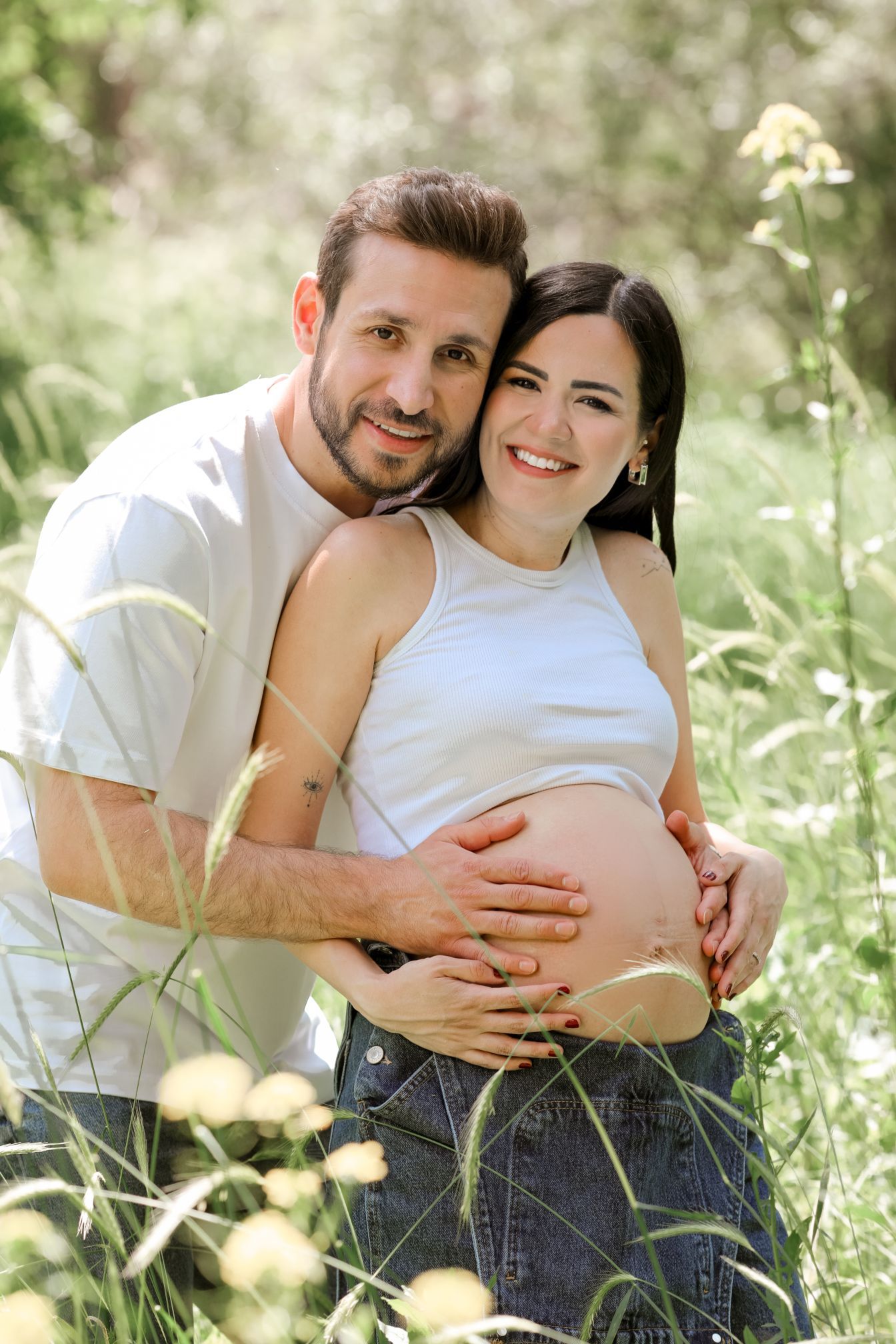 Couple embracing in sunny meadow during maternity photoshoot.
