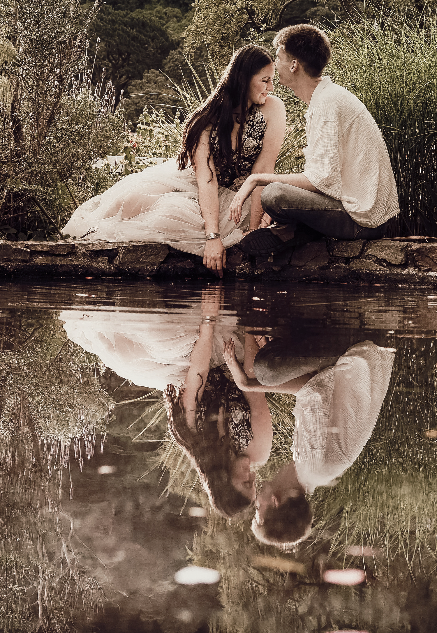 Romantic couple in love sits by reflective garden pond in beautiful outdoor engagement portrait.