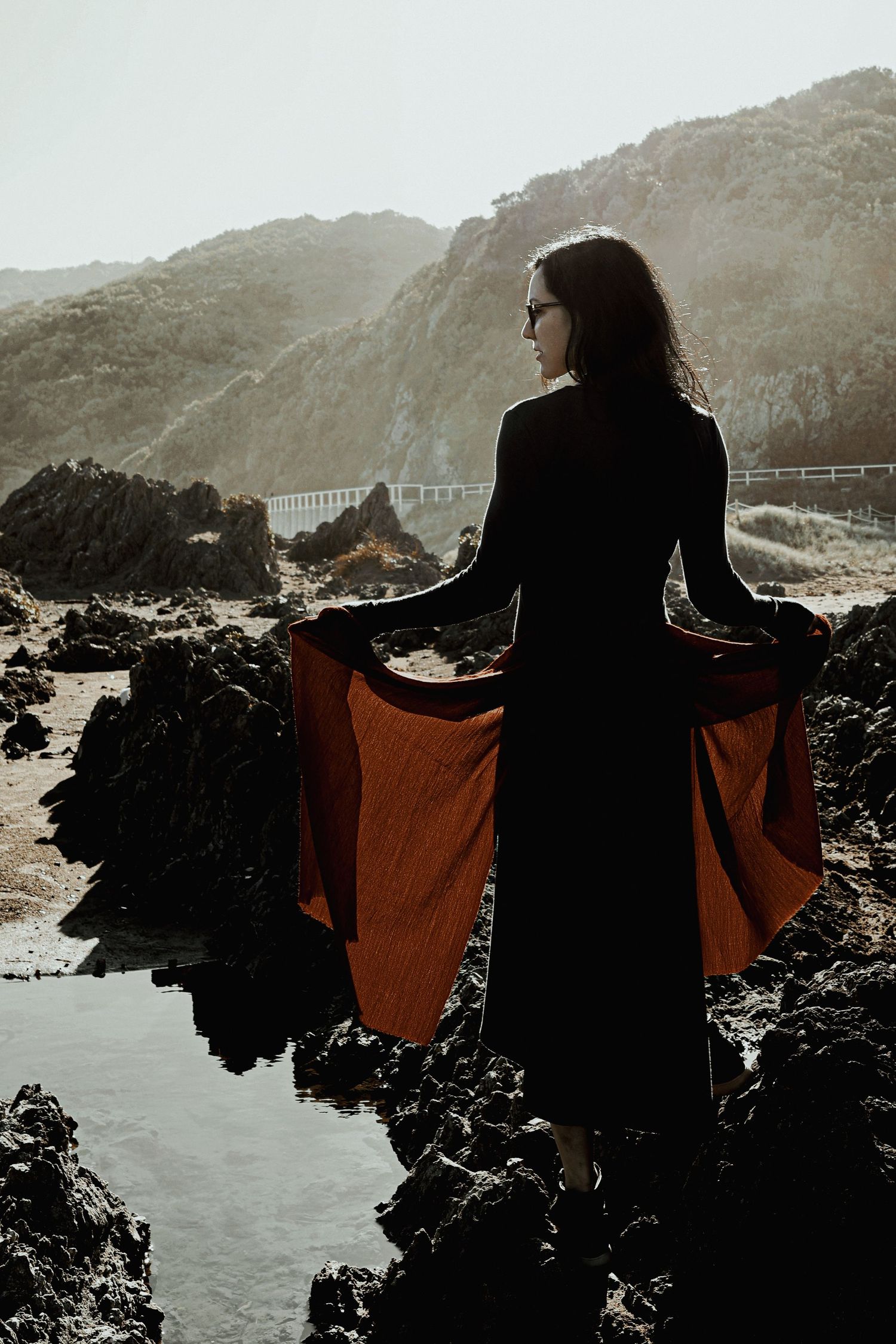 Woman in black top and rust skirt stands on rocky coastal shore, holding skirt wide with mountains behind.