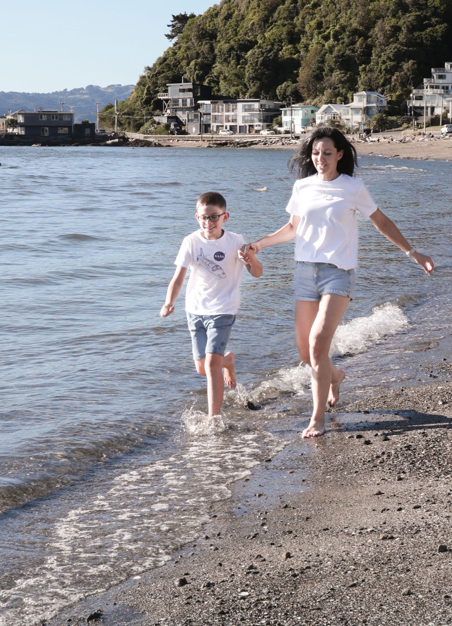 A woman and young boy hold hands running along a sandy beach shoreline with coastal houses in the background.