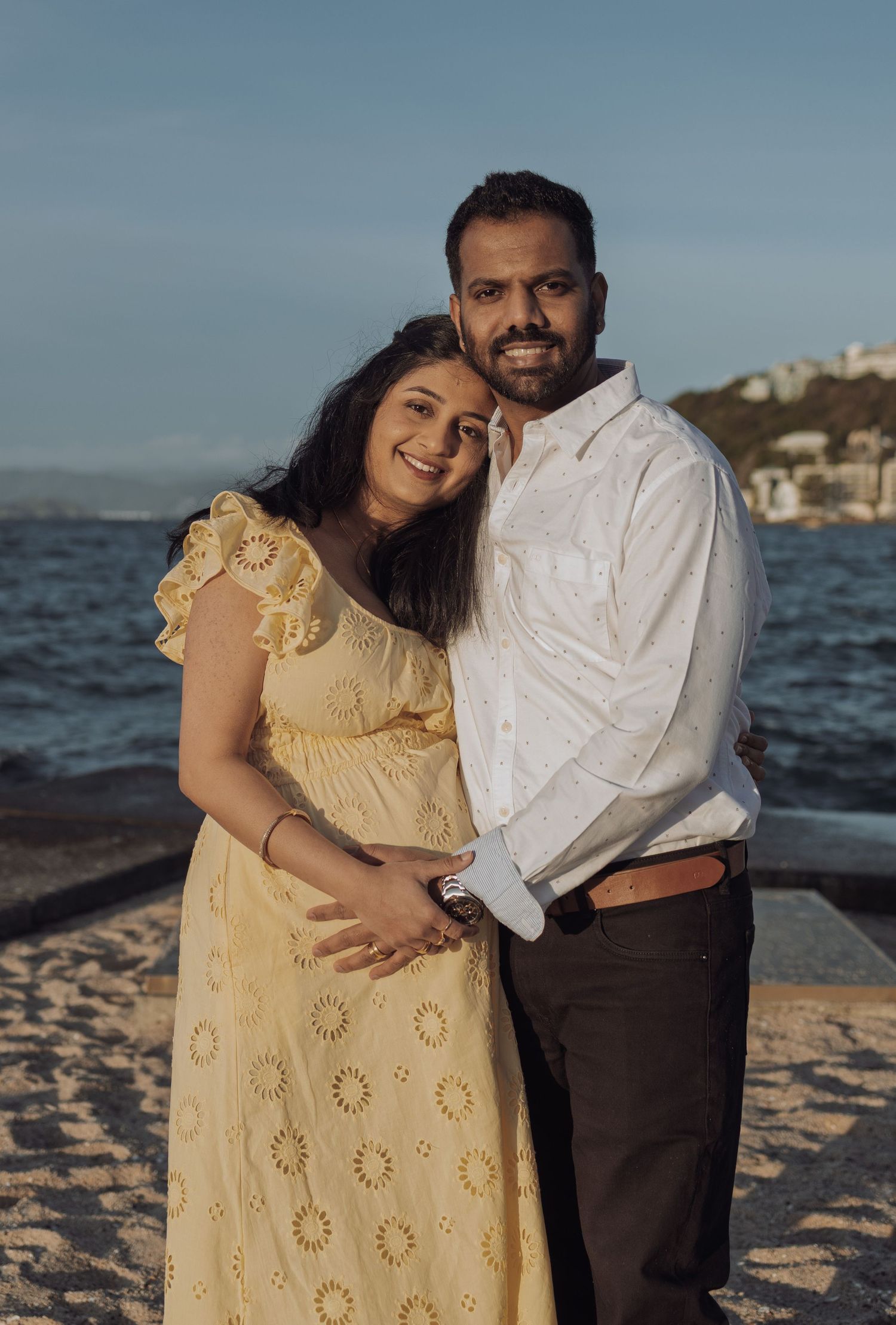 Pregnant woman in yellow dress and man in white shirt embrace smiling by the ocean on a sunny day.