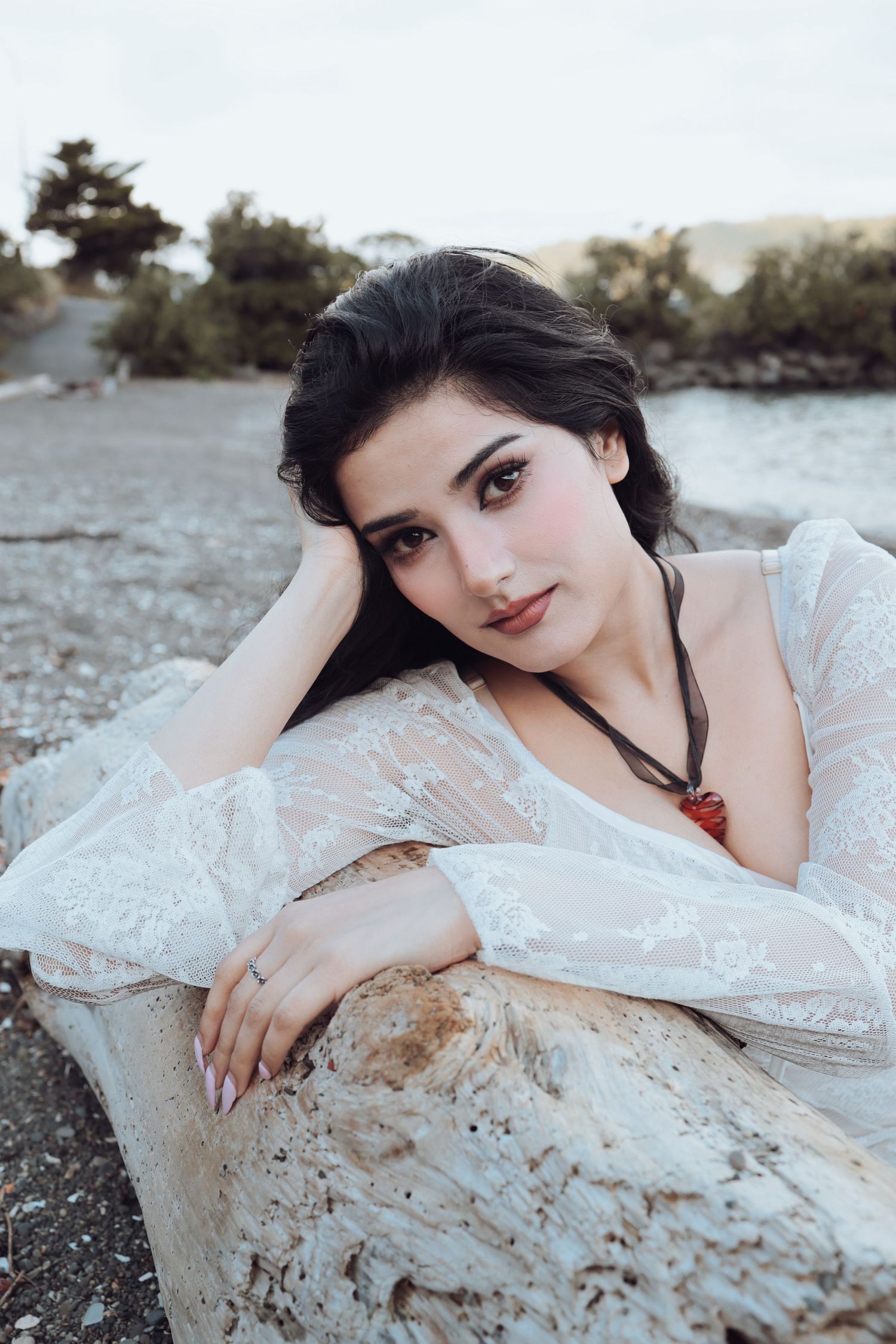Dark-haired woman in white lace dress resting on driftwood at a rocky beach.