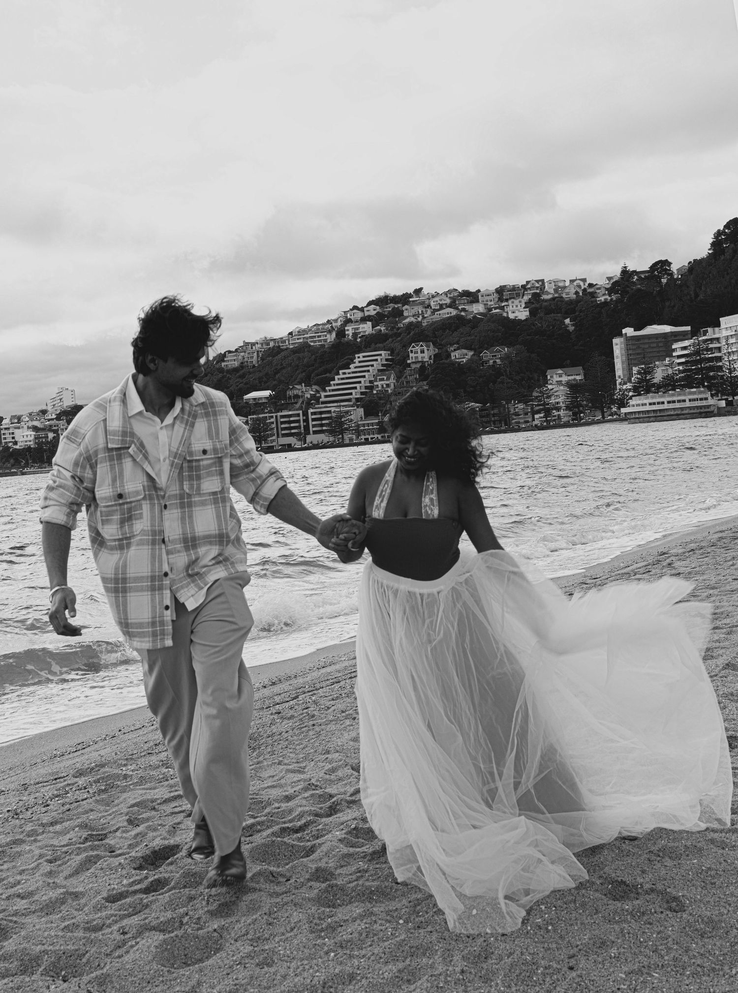 Black and white photo of a couple holding hands and walking on a beach, woman wearing a flowing white tulle skirt.