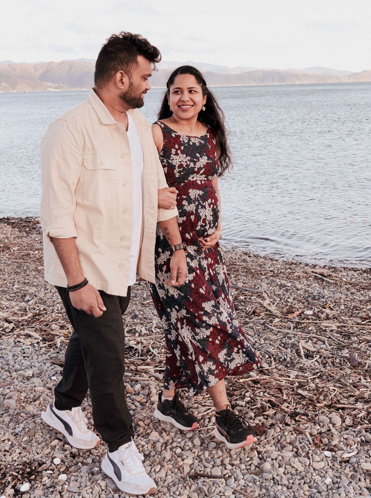 Couple posing on a rocky beach shore, woman in floral dress with baby bump, man in beige shirt.