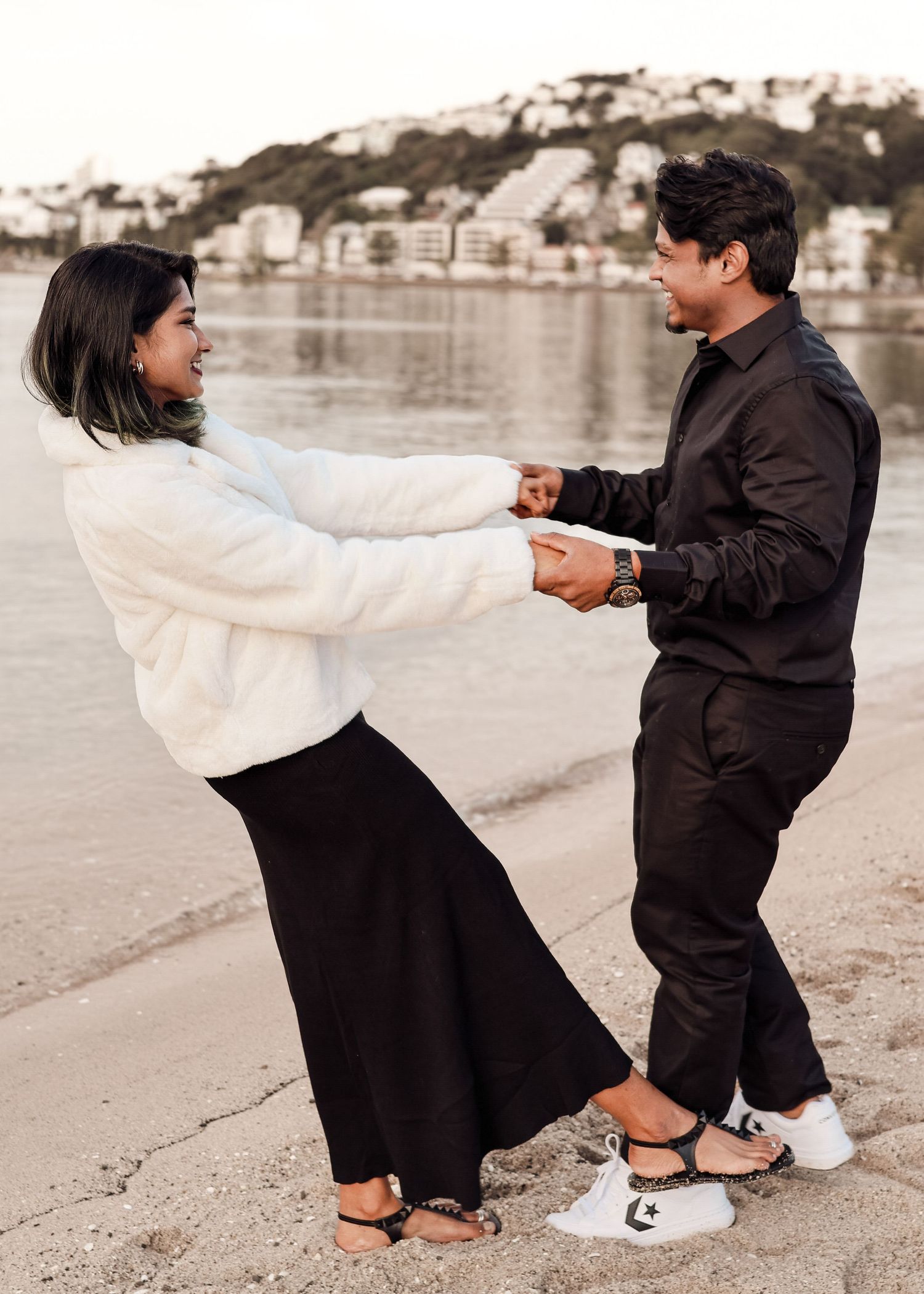 Joyful couple holding hands and dancing playfully on a sandy beach near the water.