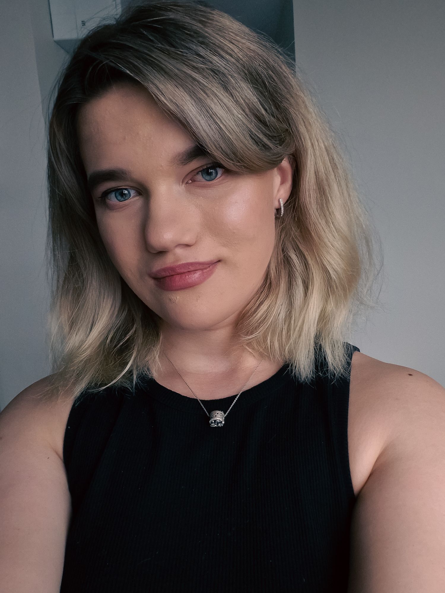 Young woman with blonde bob hair and blue eyes wearing a black sleeveless top and delicate necklace, smiling softly.