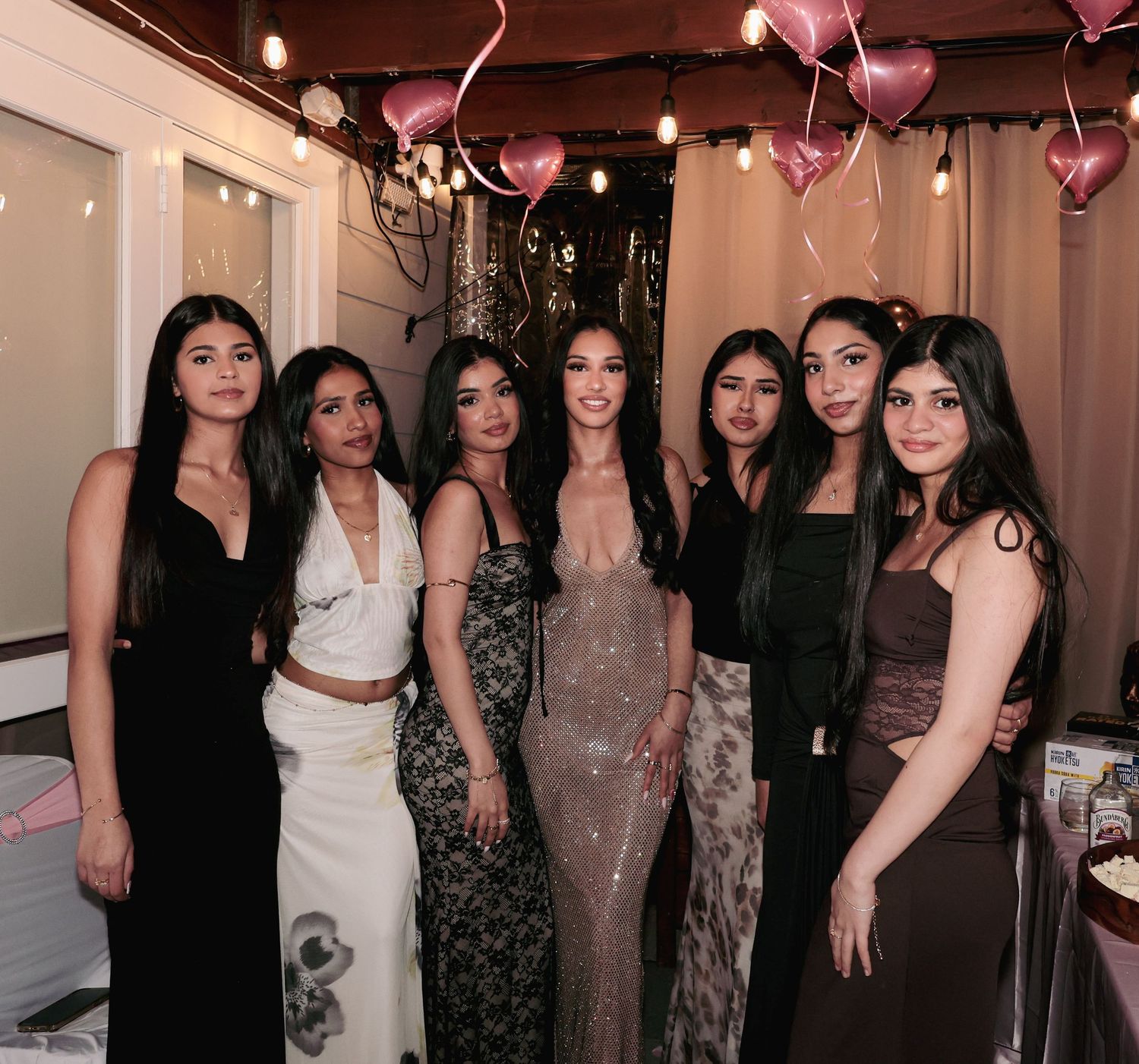 Group of seven women posing together at a party with pink heart balloons in the background.