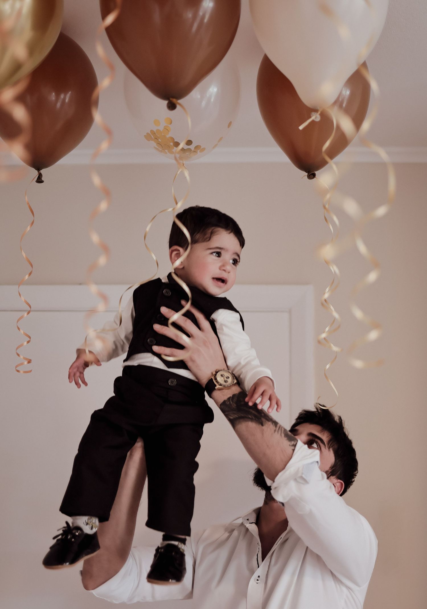 Father lifting baby boy dressed in formal suit under brown and white balloons at a party.