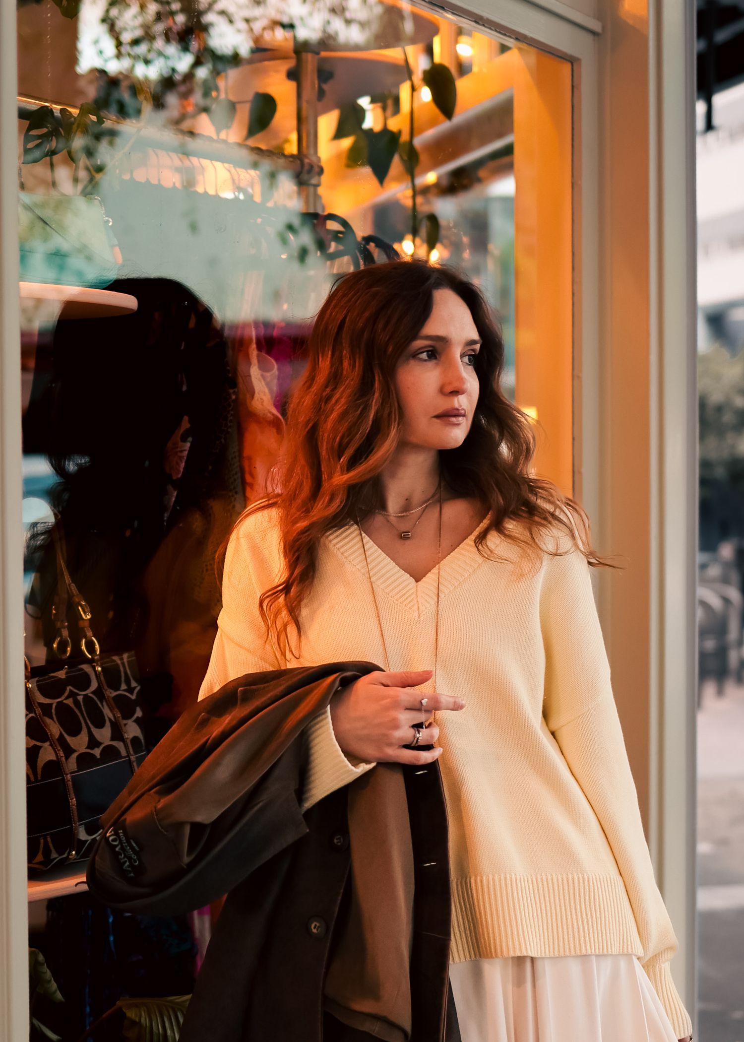 Stylish woman in cream sweater and white skirt standing outside a boutique shop at dusk.