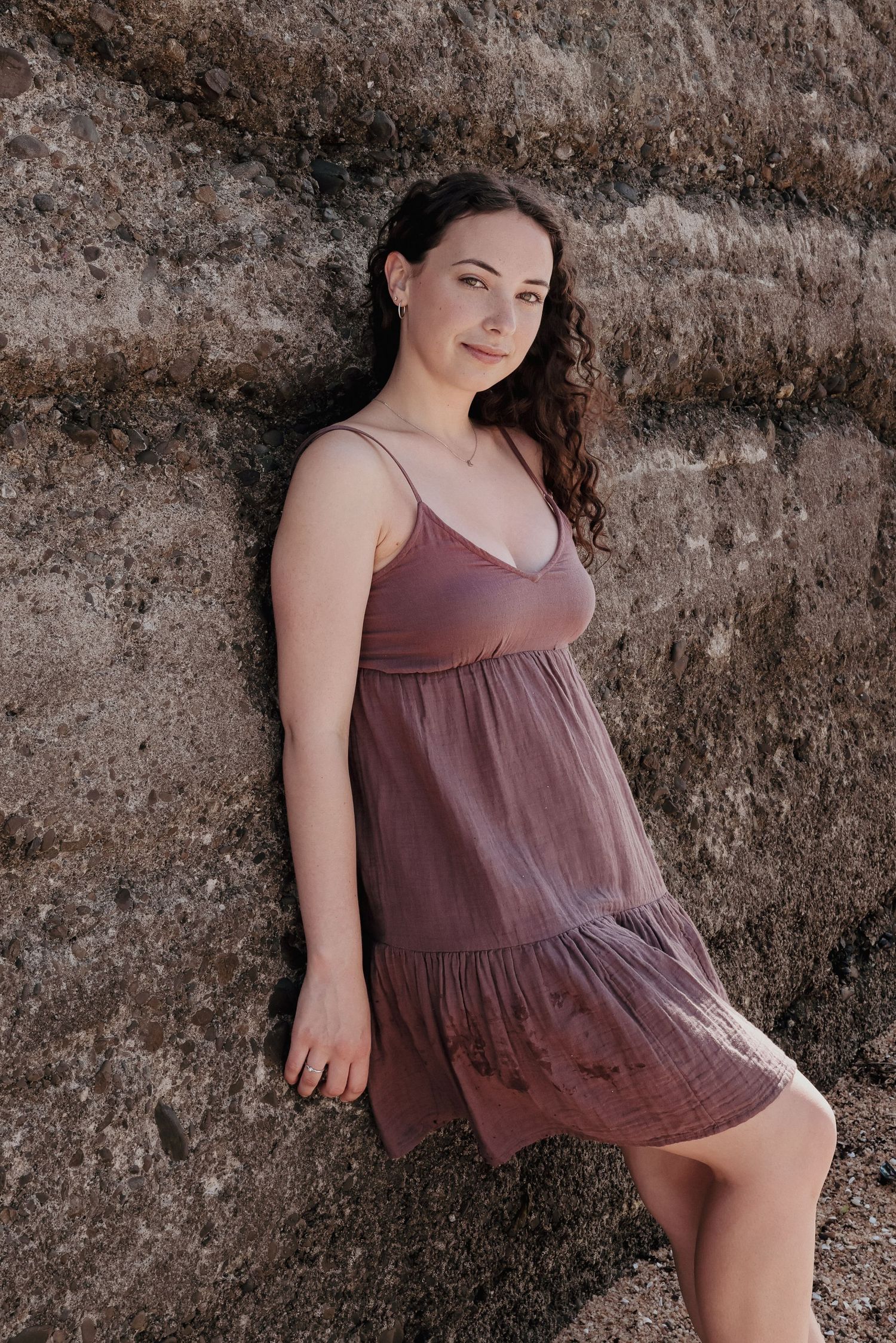 Brunette woman in mauve sundress leaning against a textured rock wall.