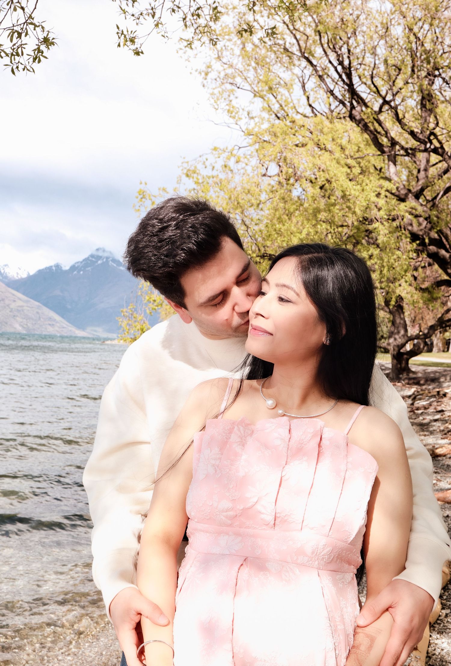 Couple sharing a tender kiss by a scenic lake with snow-capped mountains, woman in pink dress.