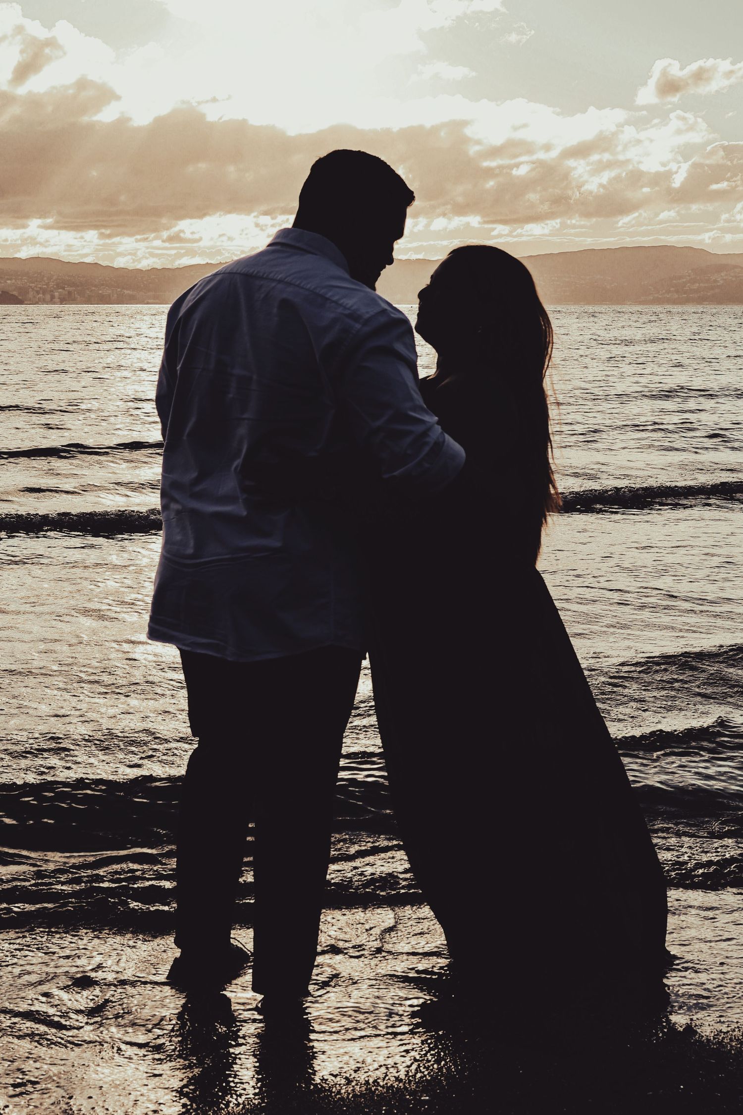 Romantic couple silhouette embracing on a beach at sunset with ocean waves and dramatic cloudy sky.