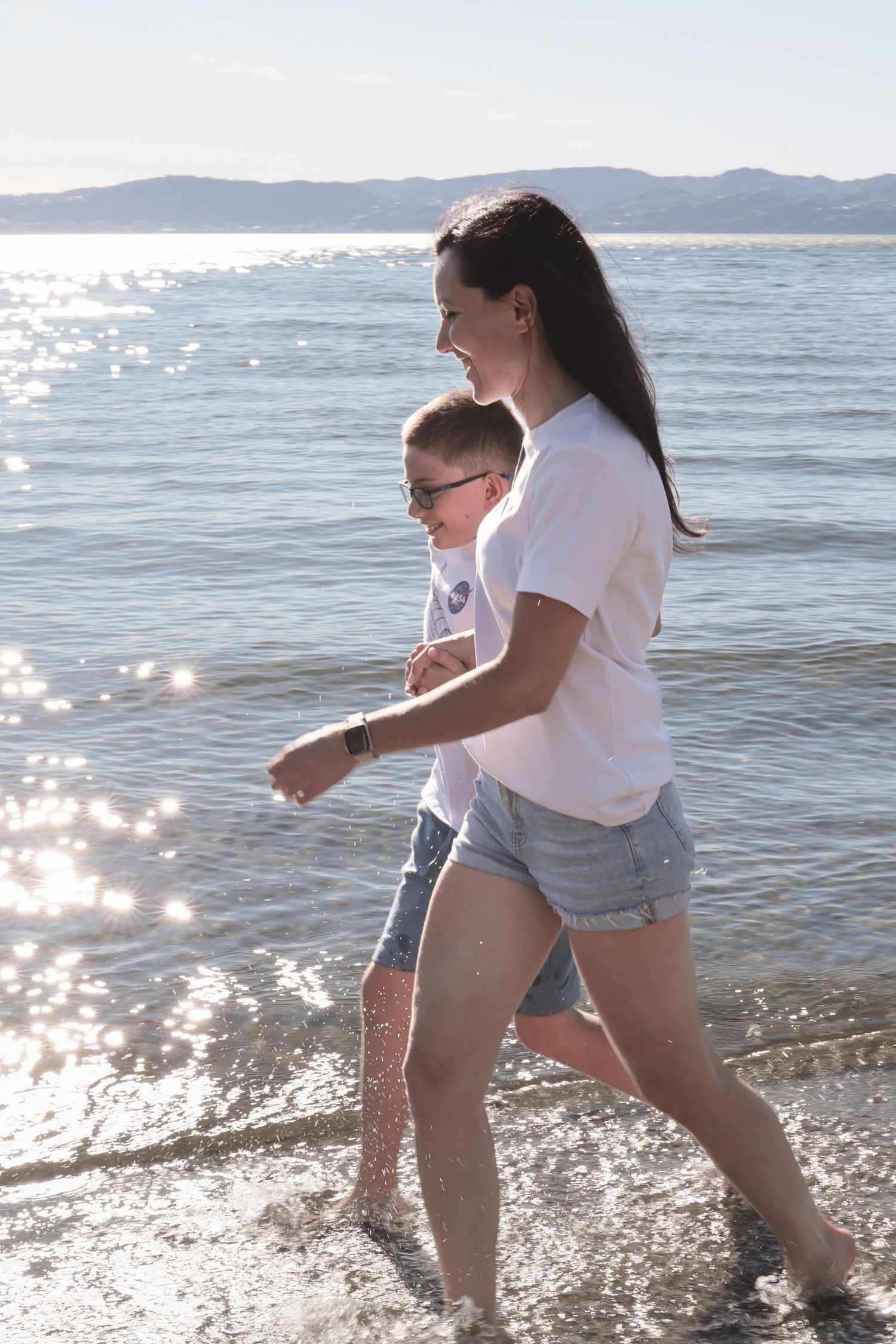 Woman and child walking along shallow beach shoreline in white shirts and denim shorts on a sunny day.
