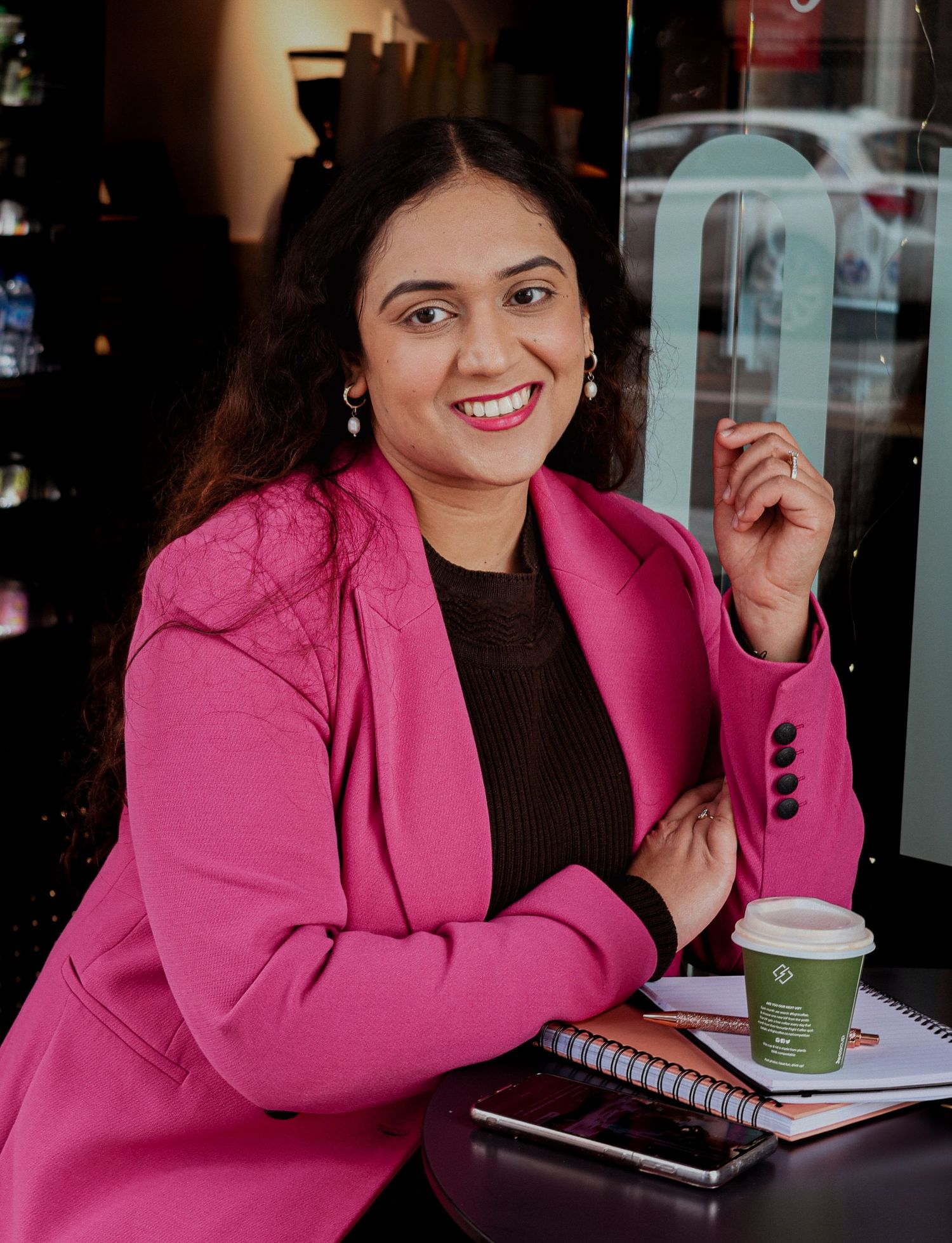 Woman in pink blazer smiling at cafe table with coffee cup and notebook.