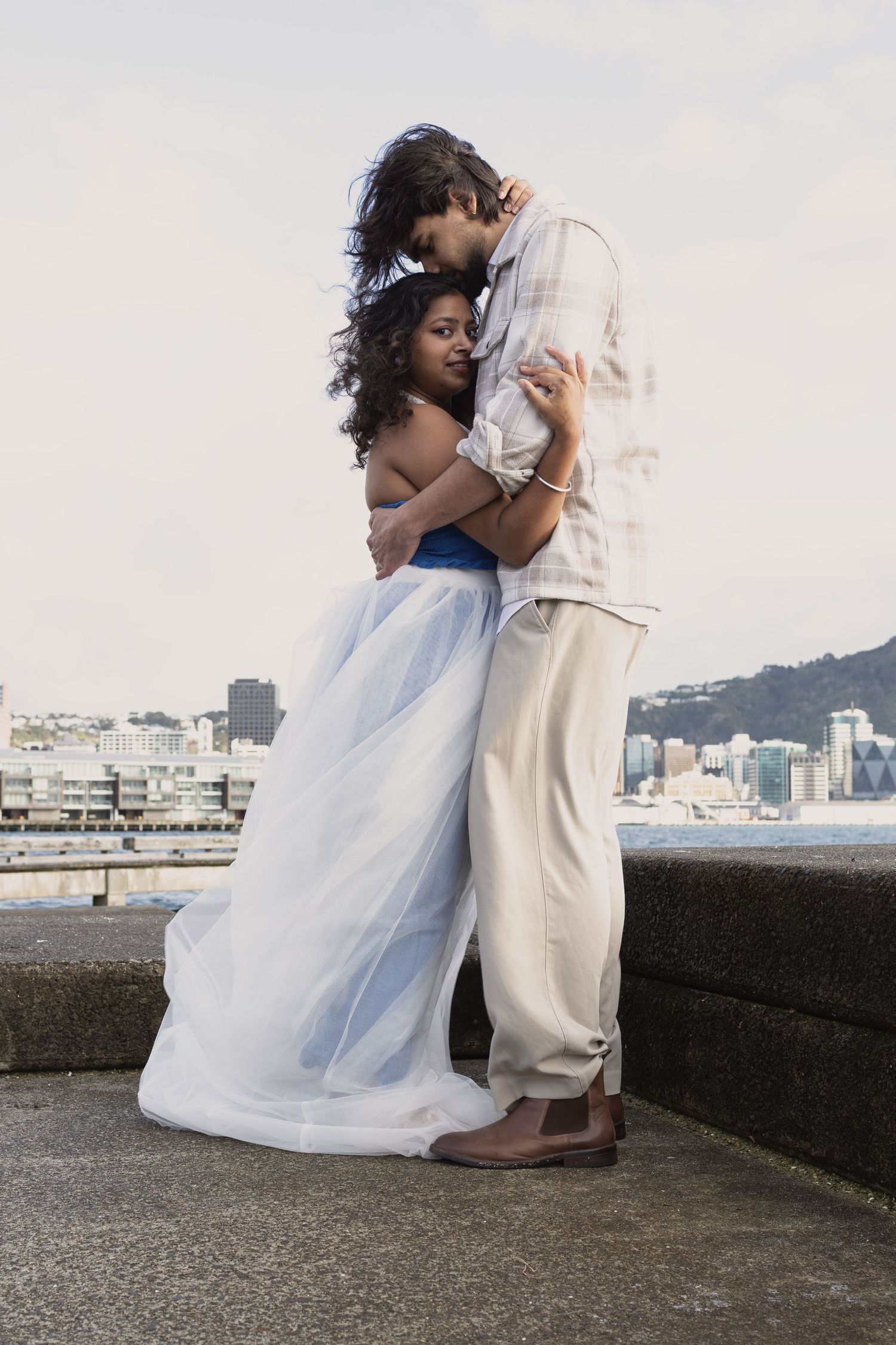 Couple embracing tenderly on a waterfront promenade, woman in blue tulle skirt, man in linen shirt, city skyline behind.