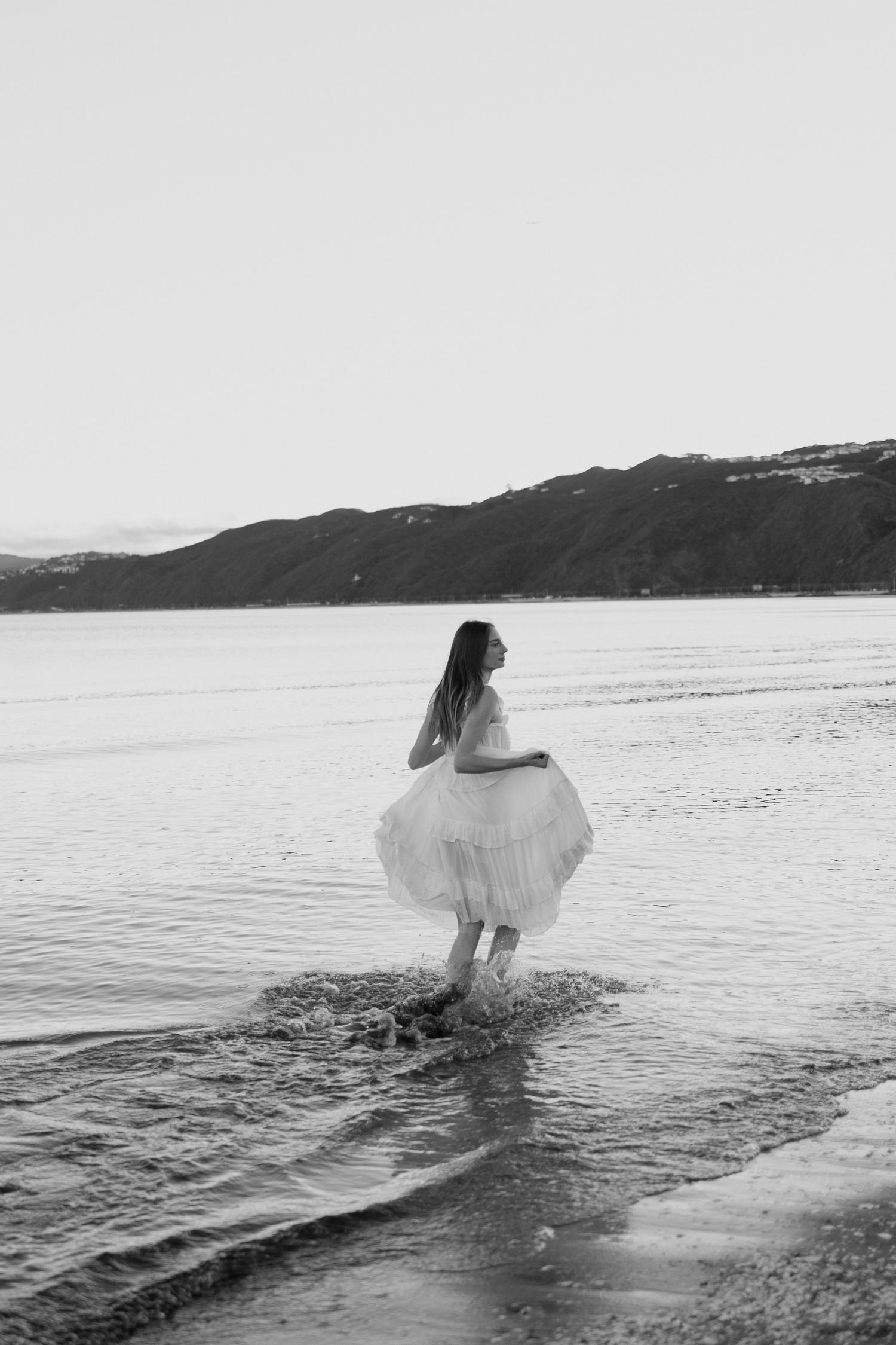 Black and white photo of a woman in a white dress wading in ocean waves with mountains in the background.
