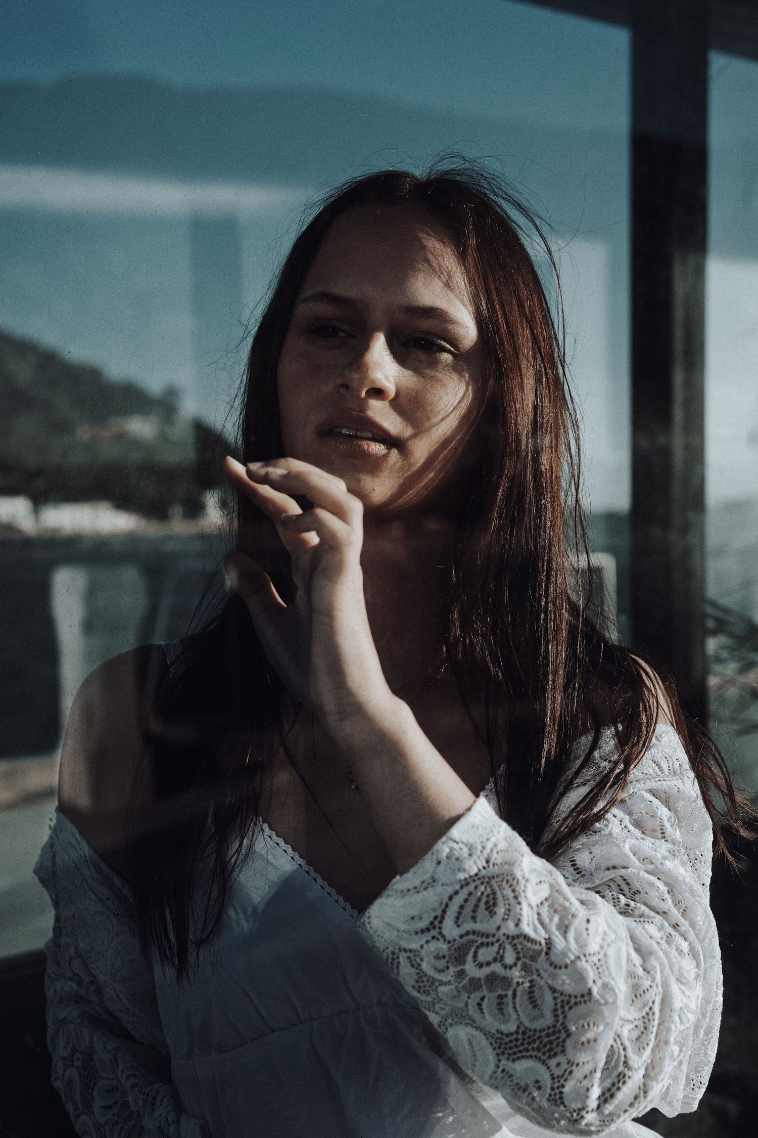 A young woman with long brown hair in a white lace top poses thoughtfully near a glass window with a scenic view.