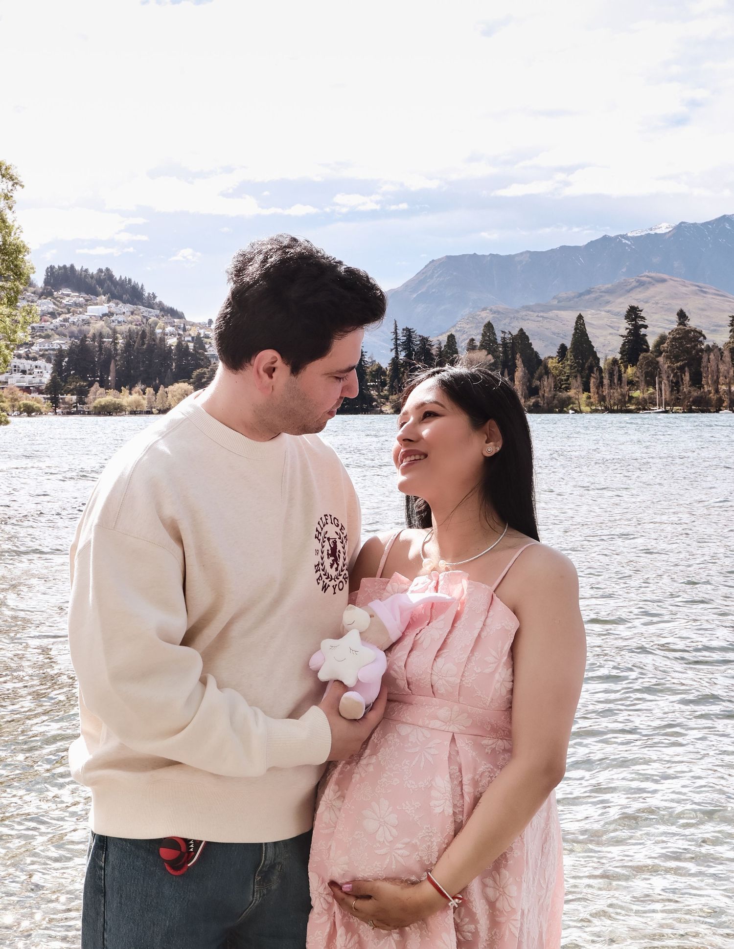 Couple standing by a lake holding a baby toy, woman in pink dress touching her baby bump with mountains in background.