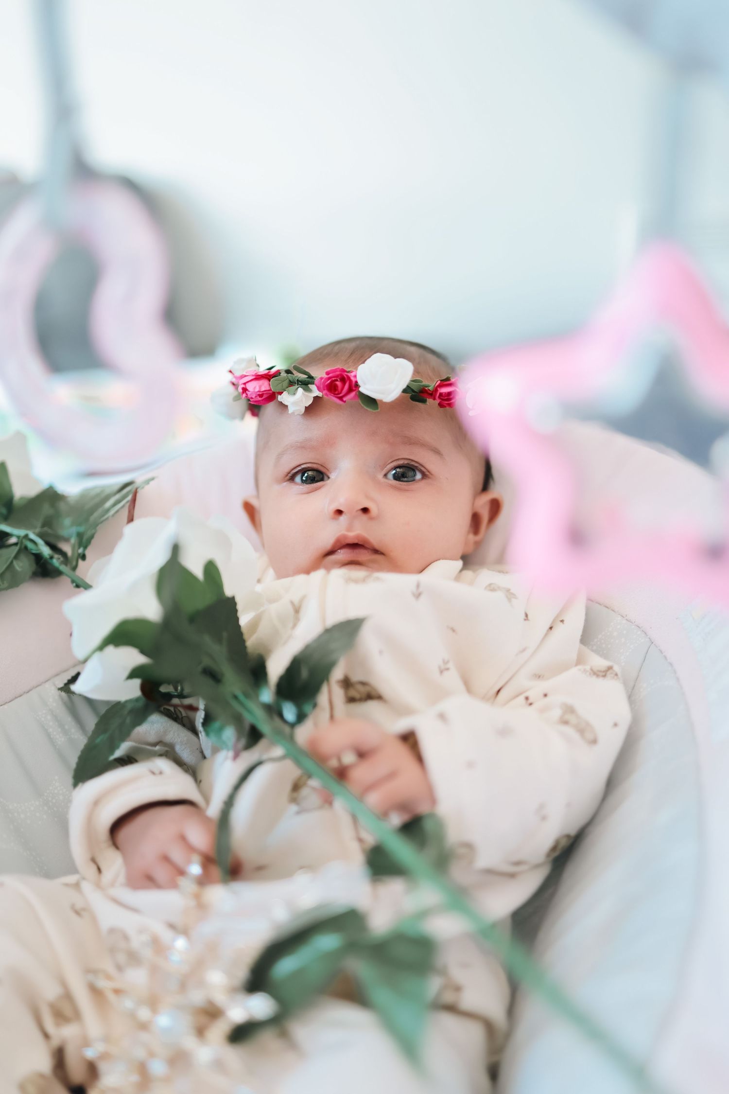 Adorable baby girl wearing a floral crown with pink and white roses, lying in a bouncer chair holding a white flower.