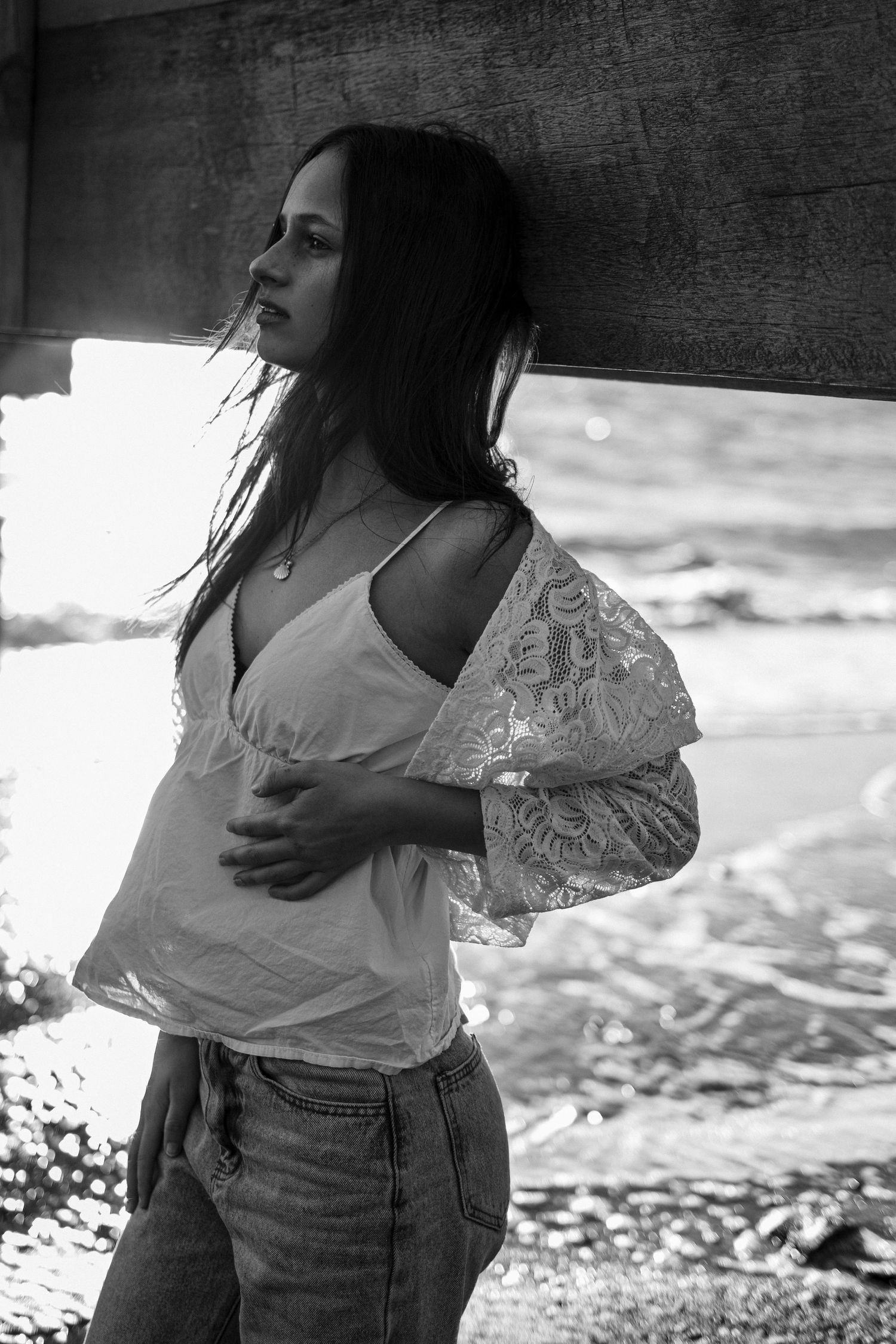 Black and white photo of a young woman in lace top and jeans leaning against a wooden post near the ocean.