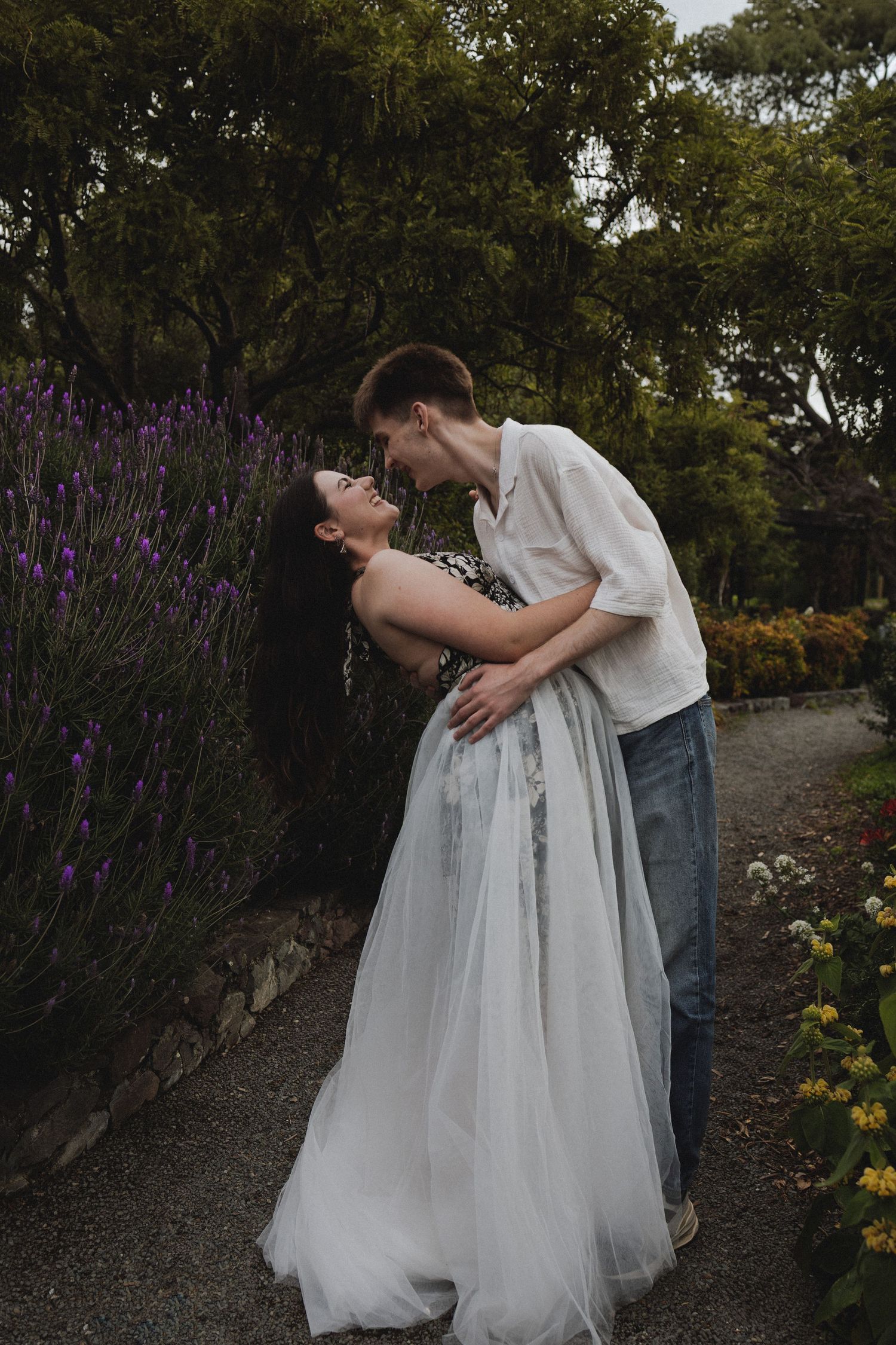 Couple sharing a romantic kiss in a garden with lavender flowers, woman in white tulle gown, lush green trees behind.