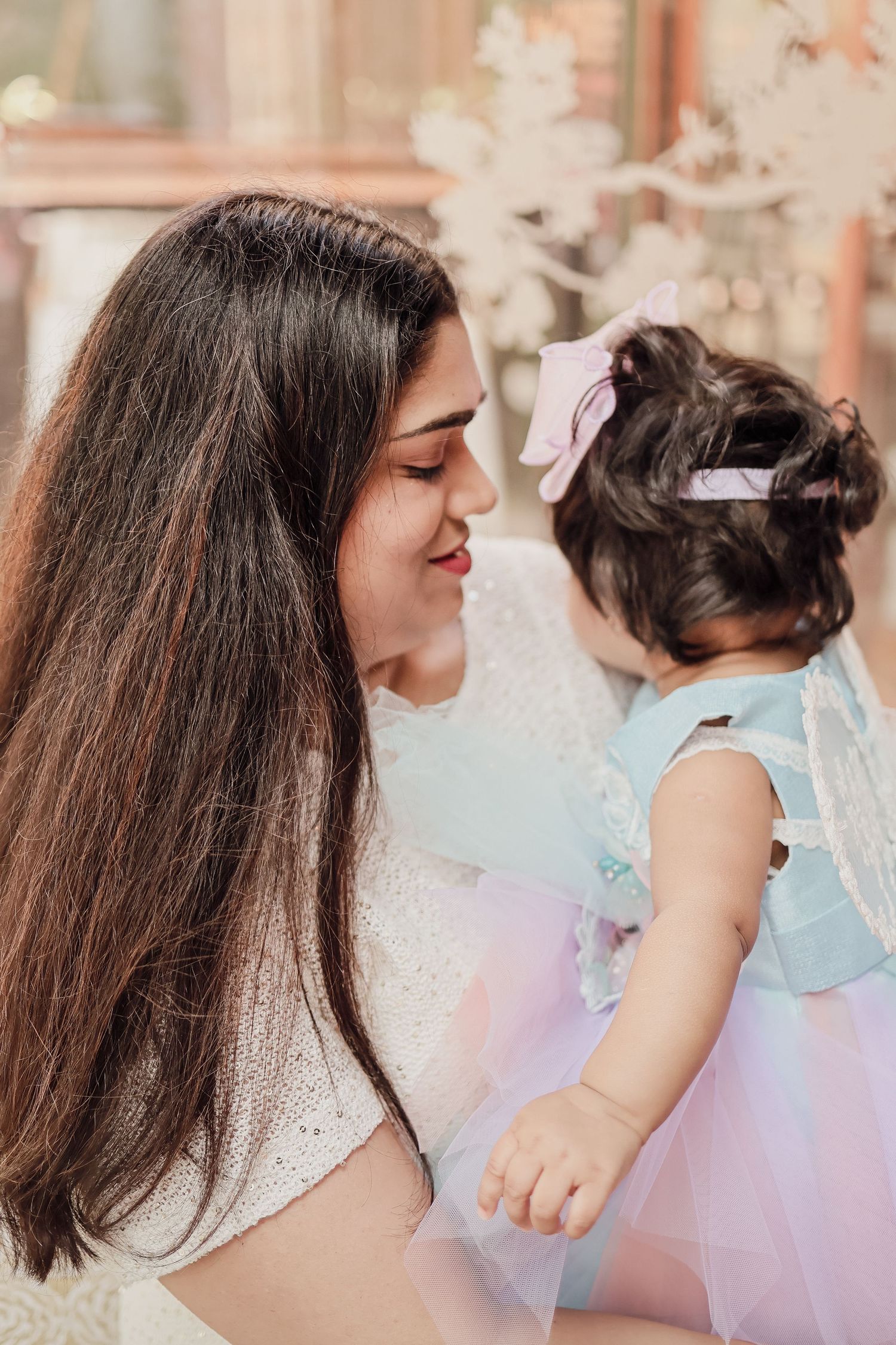 Mother tenderly touching noses with her baby daughter in a pastel tutu dress indoors.