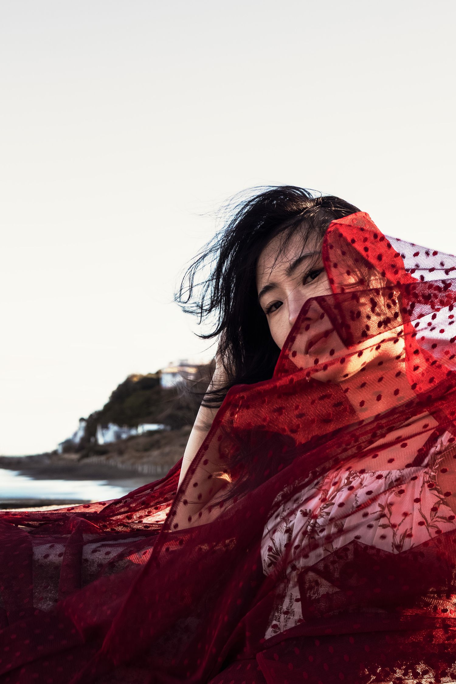 A woman with dark hair peers over a flowing red lace fabric billowing in the wind by a coastal backdrop.