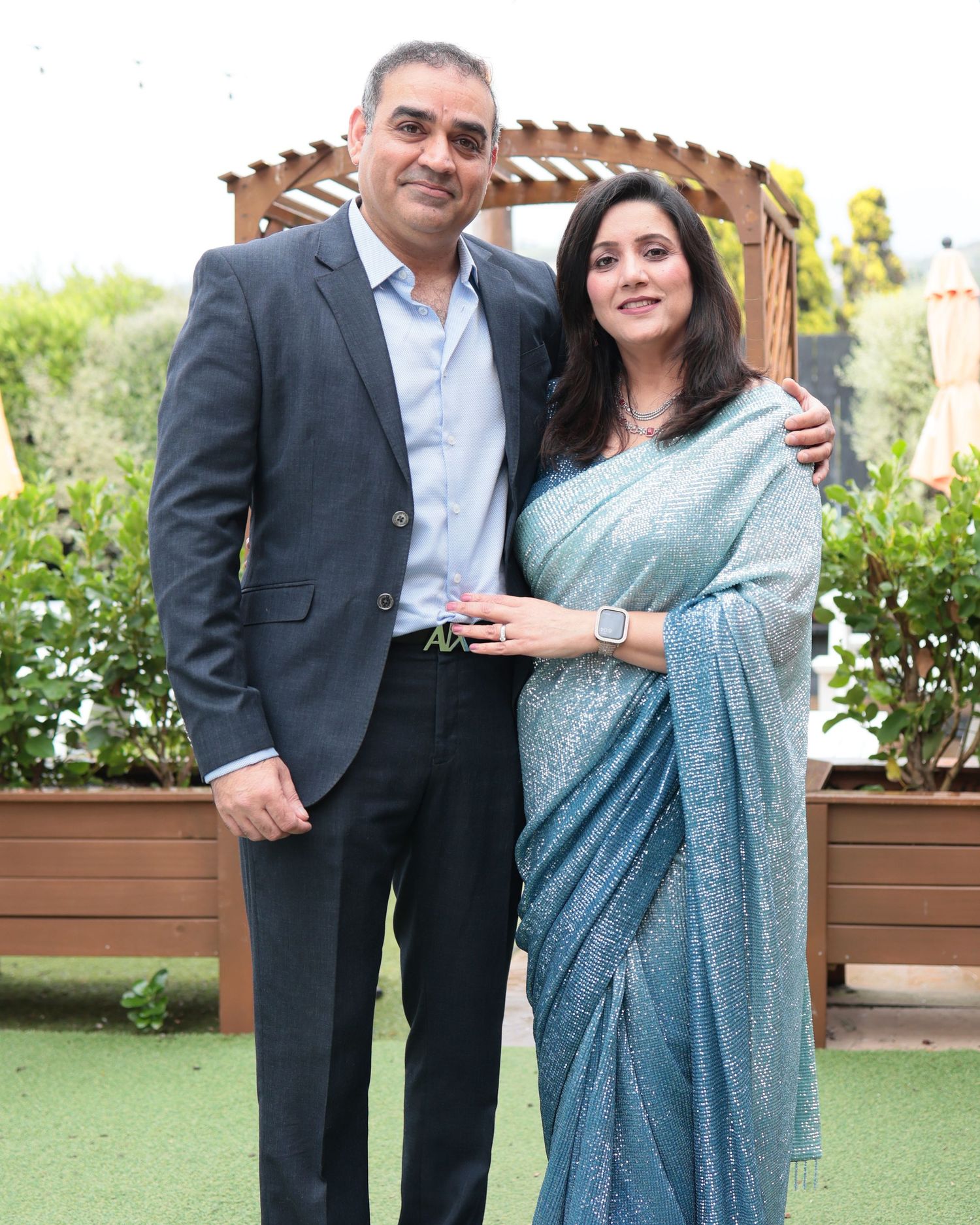 Couple posing outdoors, man in suit and woman in blue sparkly saree at a celebration event.