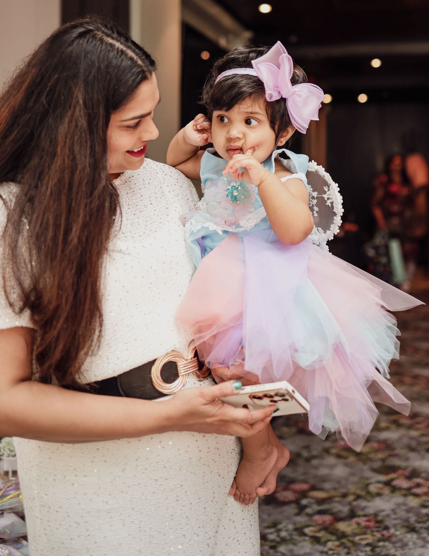 Mother in white dress holding baby girl in colorful tutu fairy costume at a birthday celebration.