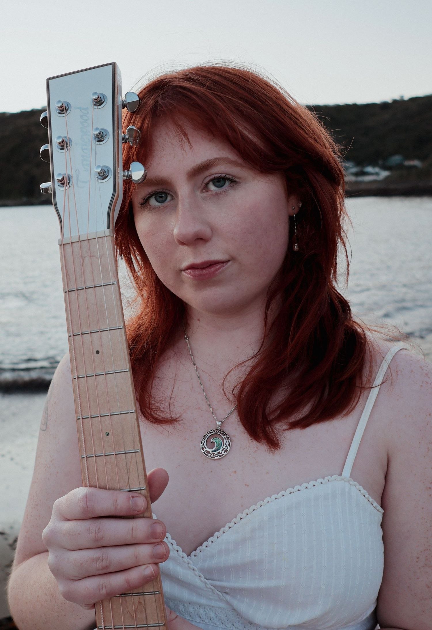 A red-haired woman holds a guitar neck while posing near a coastal waterfront at dusk.