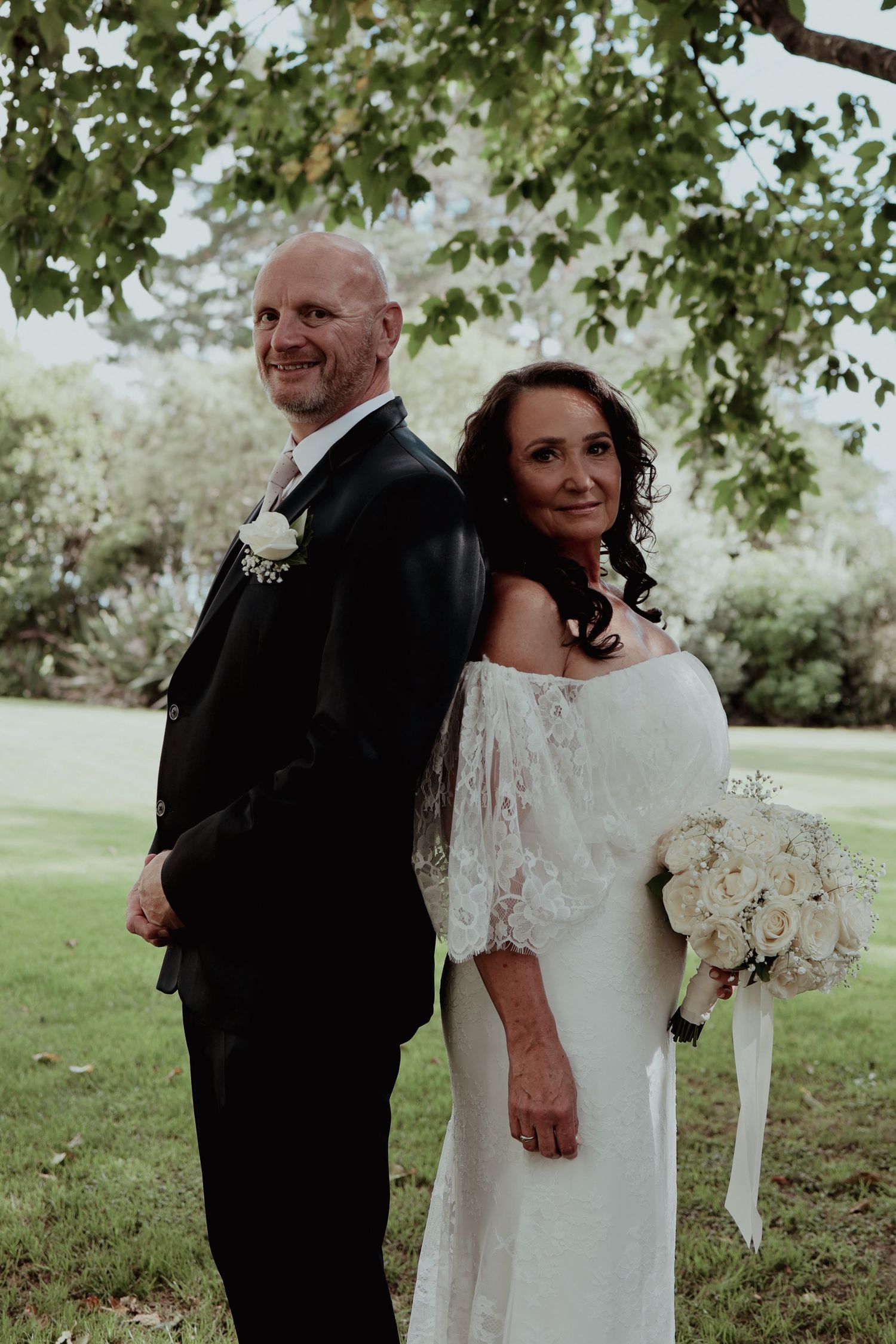 Bride and groom pose back-to-back smiling under leafy tree with white rose bouquet.