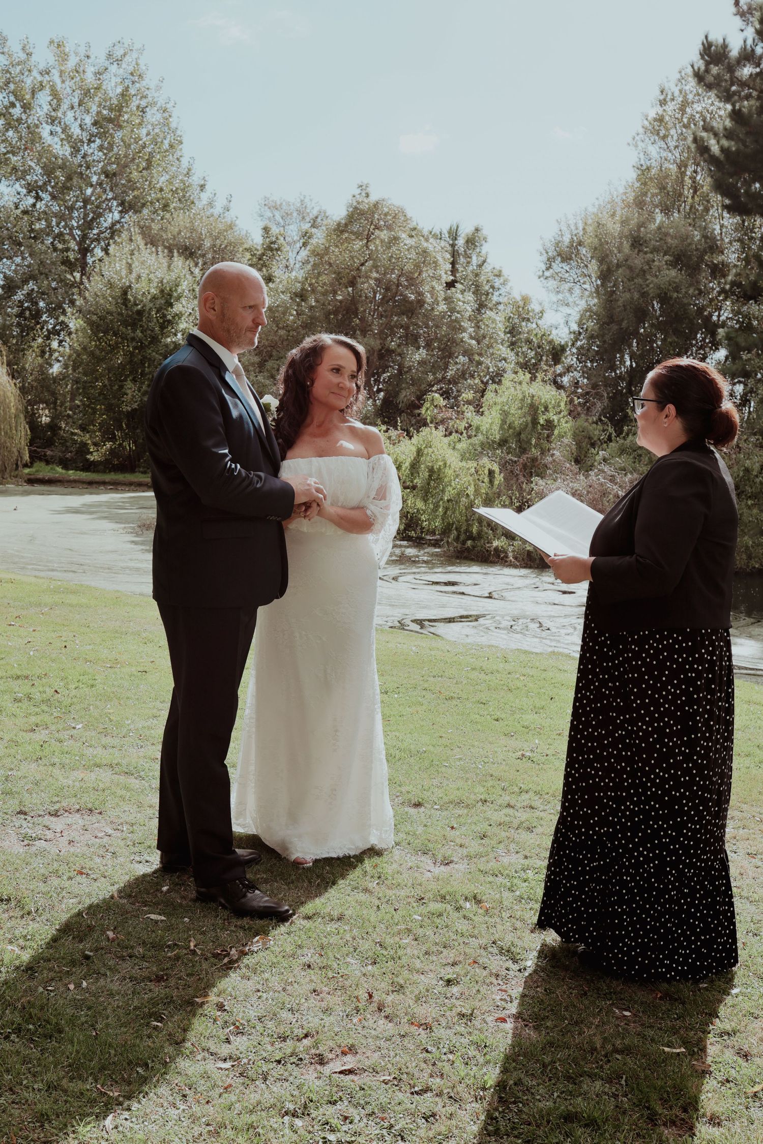 Outdoor wedding ceremony with bride in white lace gown and groom in black suit beside a pond.