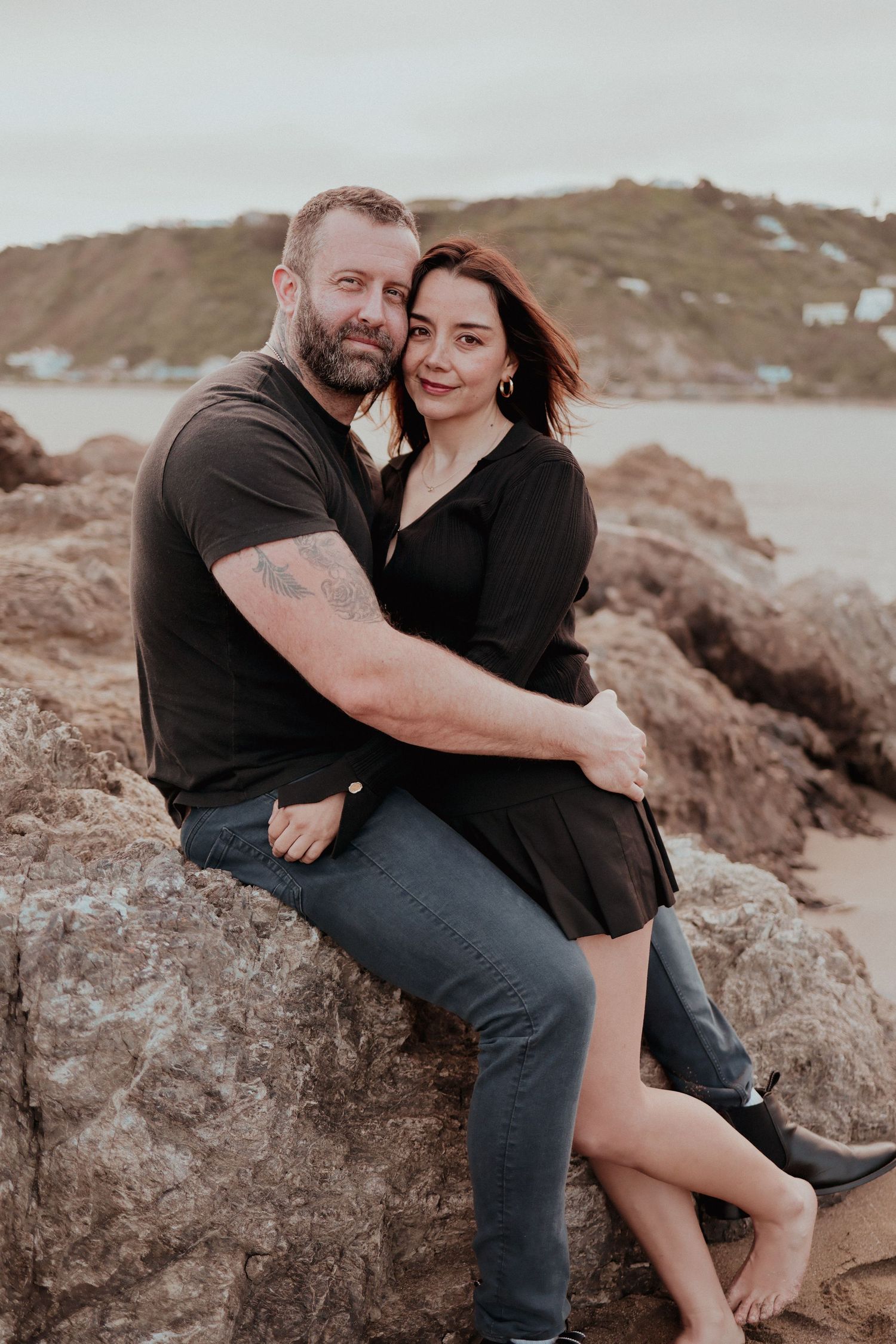 Couple in black outfits sitting on coastal rocks, embracing romantically with a scenic hillside and ocean in the background.