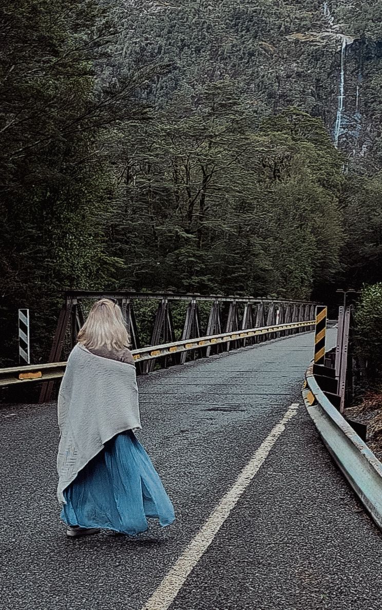 Woman in blue skirt and gray shawl walks toward a metal bridge surrounded by lush green forest and waterfall.