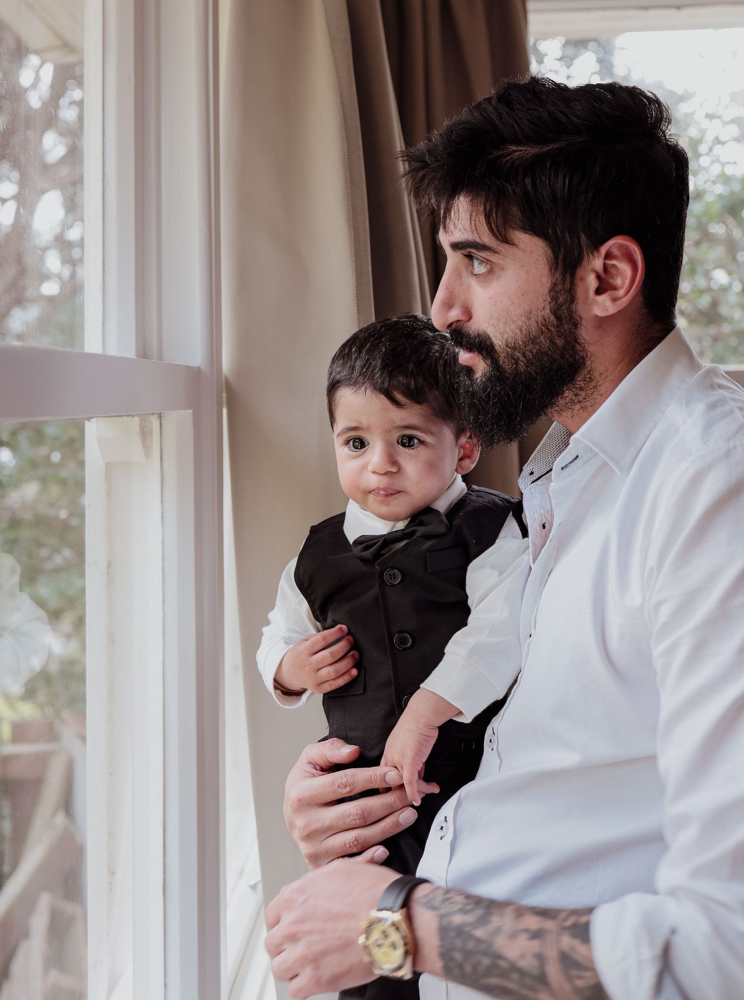 Bearded father holding baby boy in formal suit while looking out a window together.