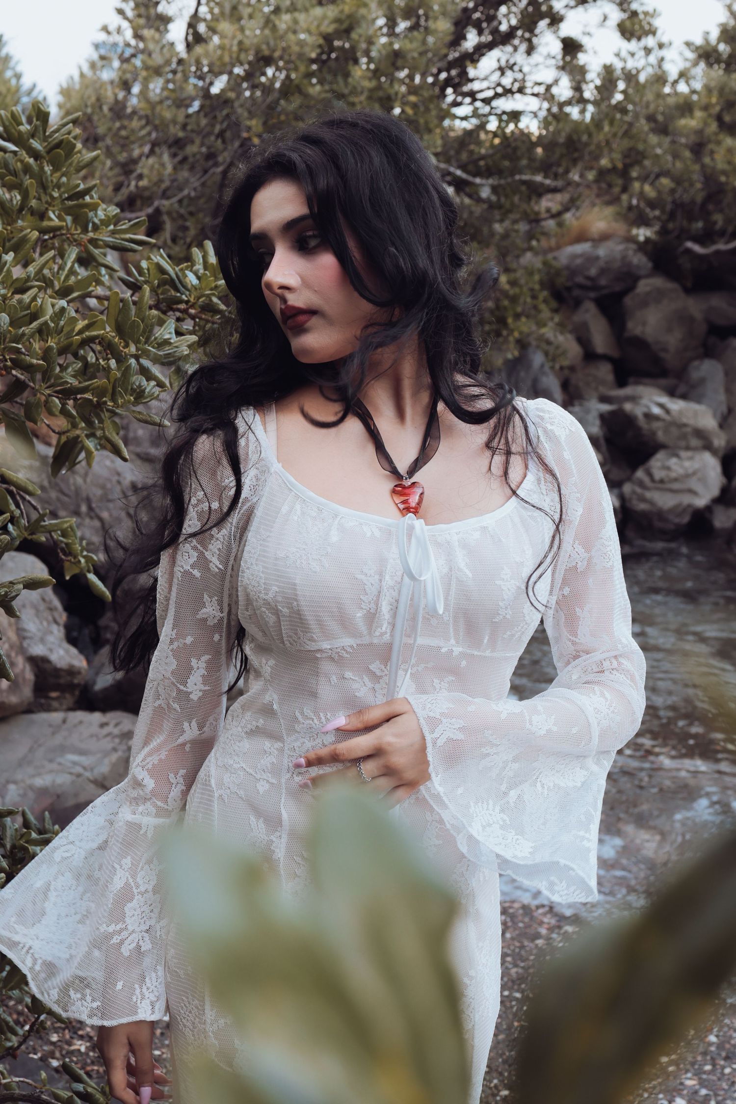 Dark-haired woman in white lace dress with red heart necklace among rocks.