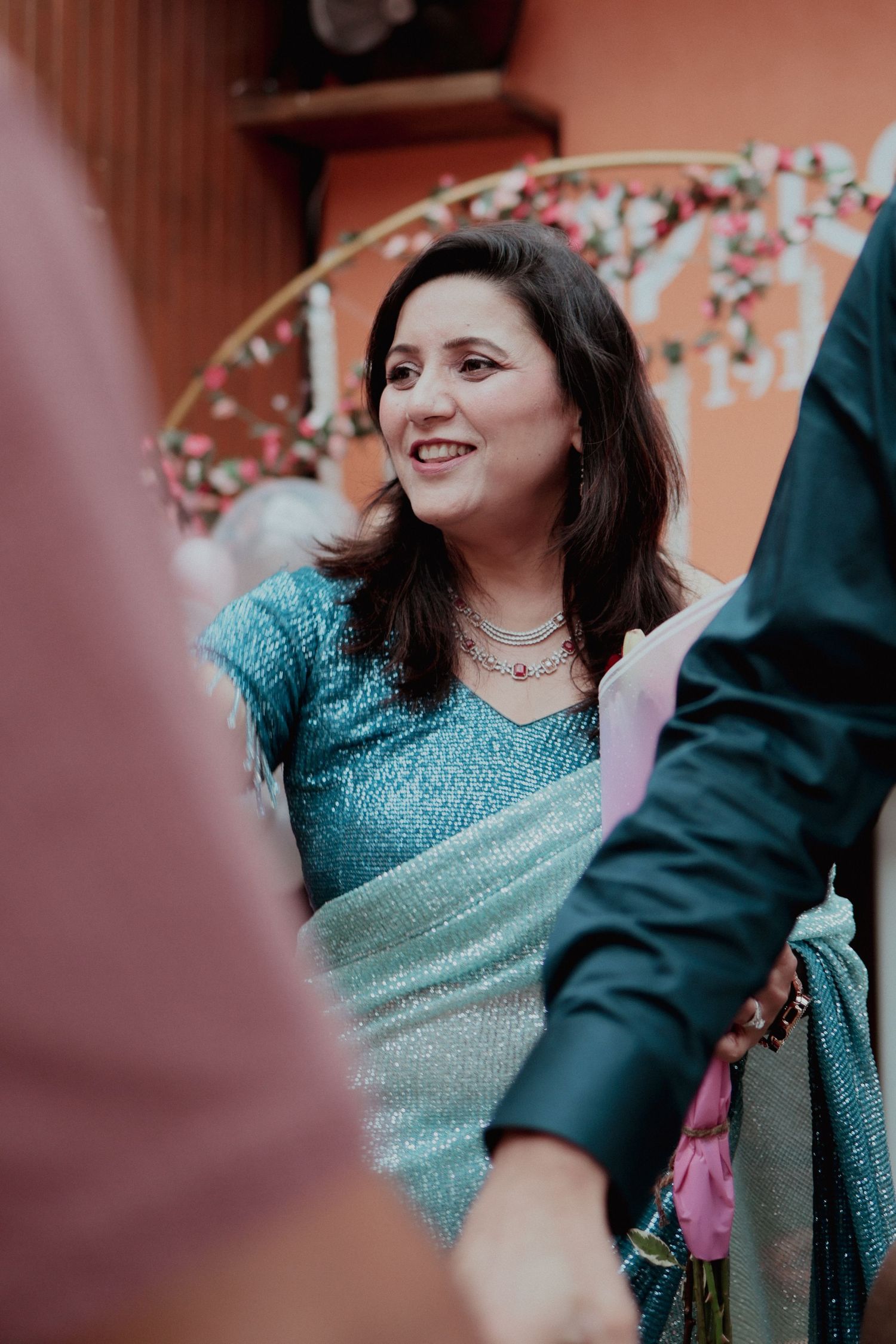 Smiling woman in teal sparkly saree at an elegant Indian celebration with floral backdrop.
