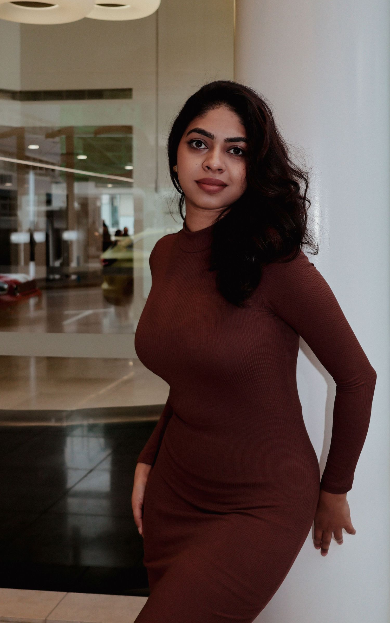 A young woman in a brown turtleneck bodycon dress poses confidently in a modern interior setting.