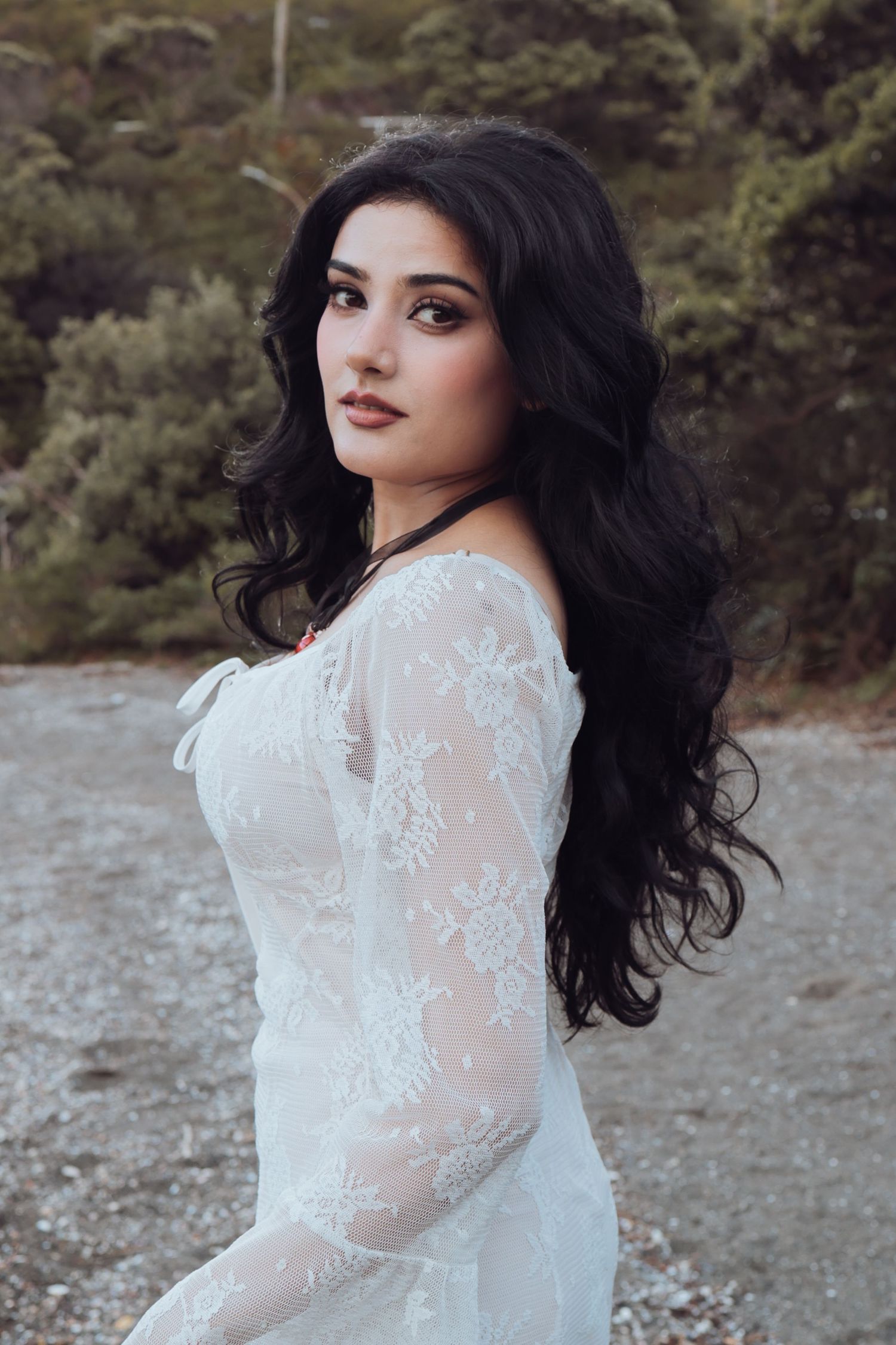A woman with long black hair wearing a white lace off-shoulder dress poses outdoors in nature.