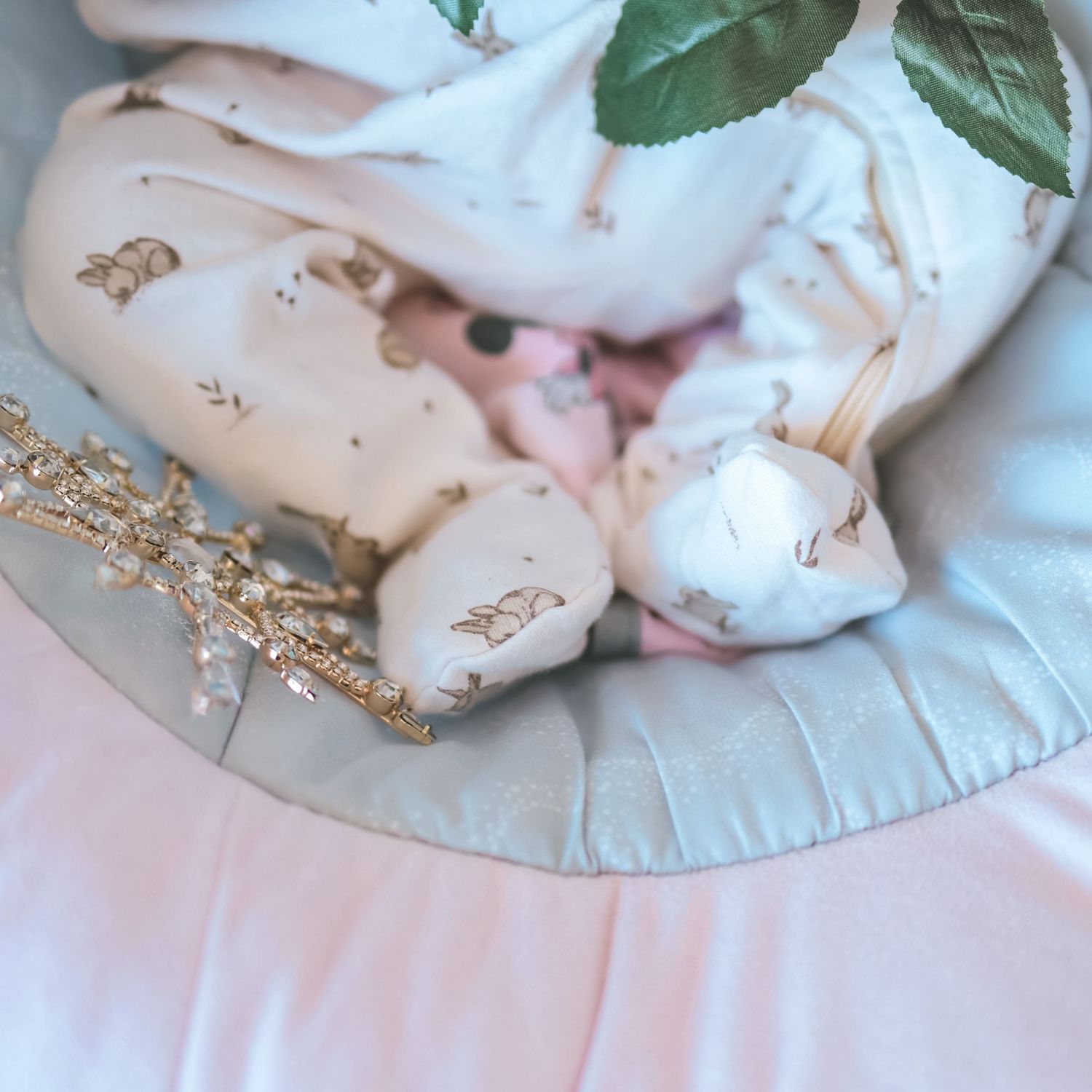A sleeping baby in a floral white onesie rests on a soft pink and sage green cushion with gold decorative branches.