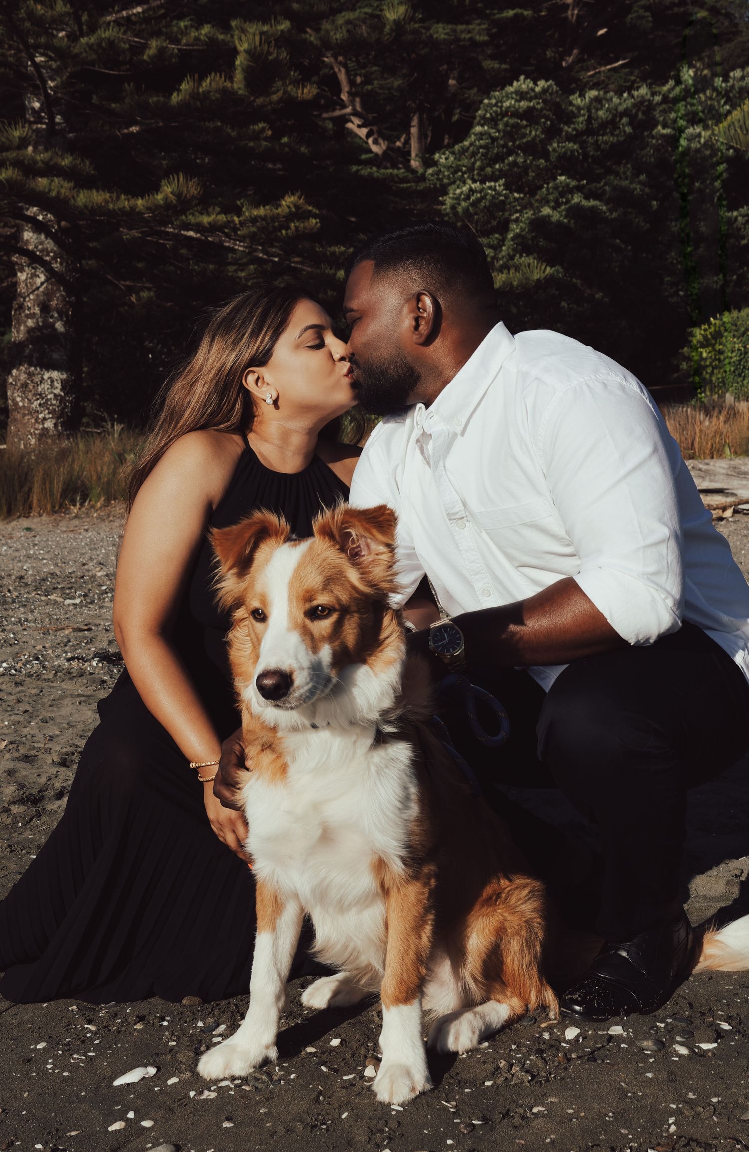 Couple kissing outdoors on a beach with their brown and white Border Collie dog sitting between them.