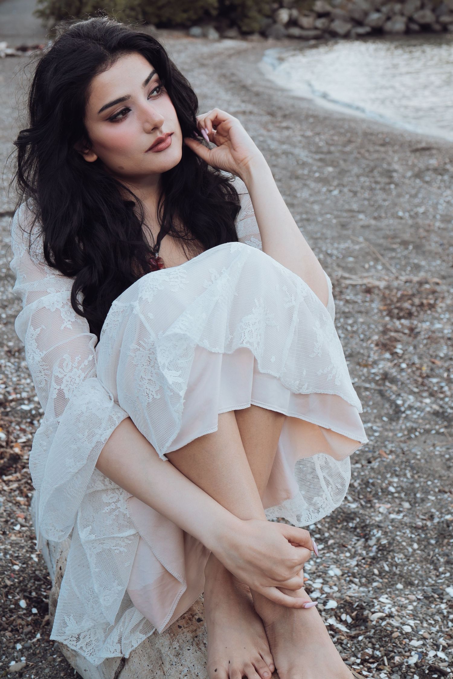 Young woman with dark hair in white lace dress sitting gracefully on rocky shore, gazing thoughtfully into distance.