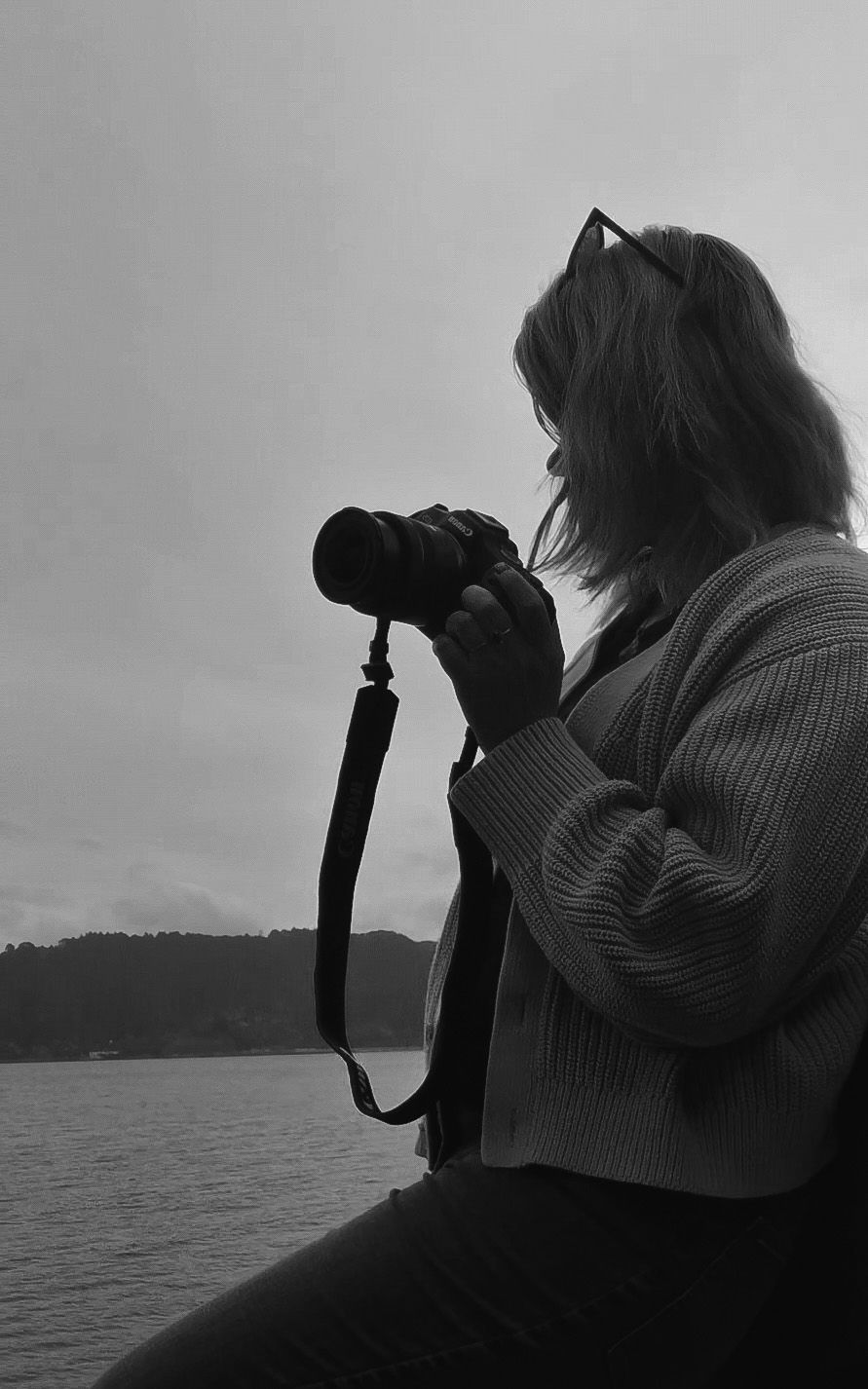 Black and white photo of a woman holding a camera by a lake, with hills in the background on a cloudy day.