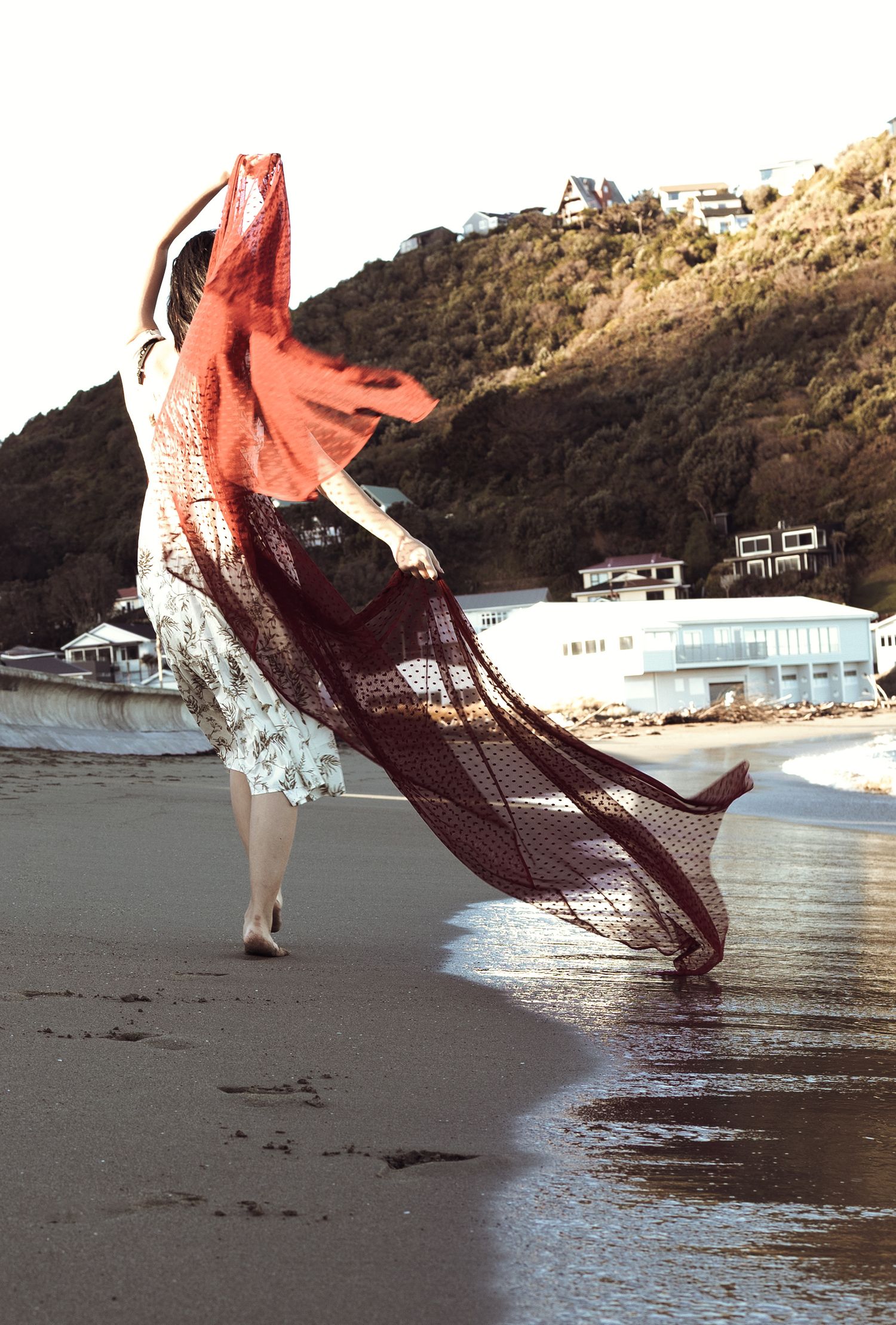 Woman in floral dress walks on beach holding flowing red mesh fabric, with hillside houses in background.