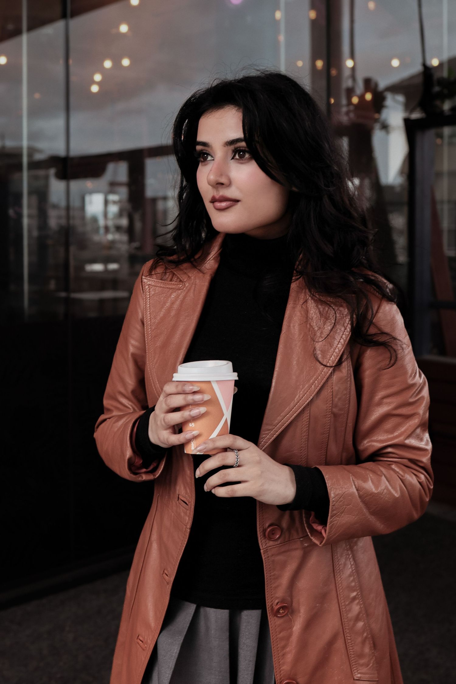 A stylish brunette woman in a rose leather trench coat holds a coffee cup outdoors near a glass building.