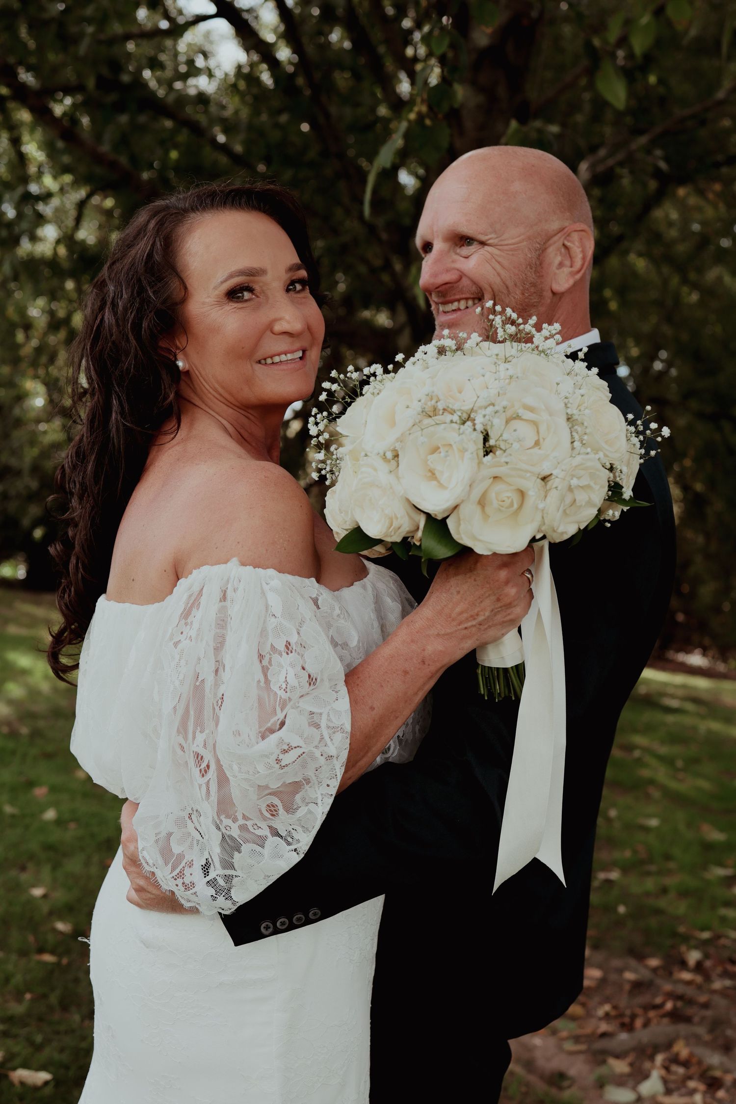 Smiling couple holds white rose and baby's breath bouquet together in lush garden.