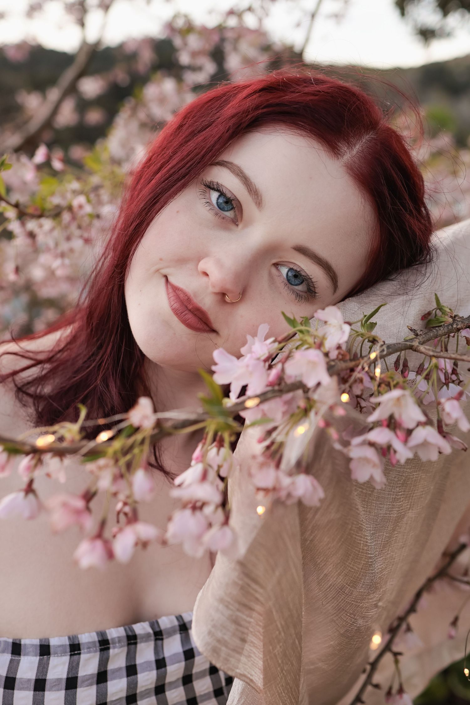 Red-haired blue-eyed woman posing among pink cherry blossom branches in a gingham top.
