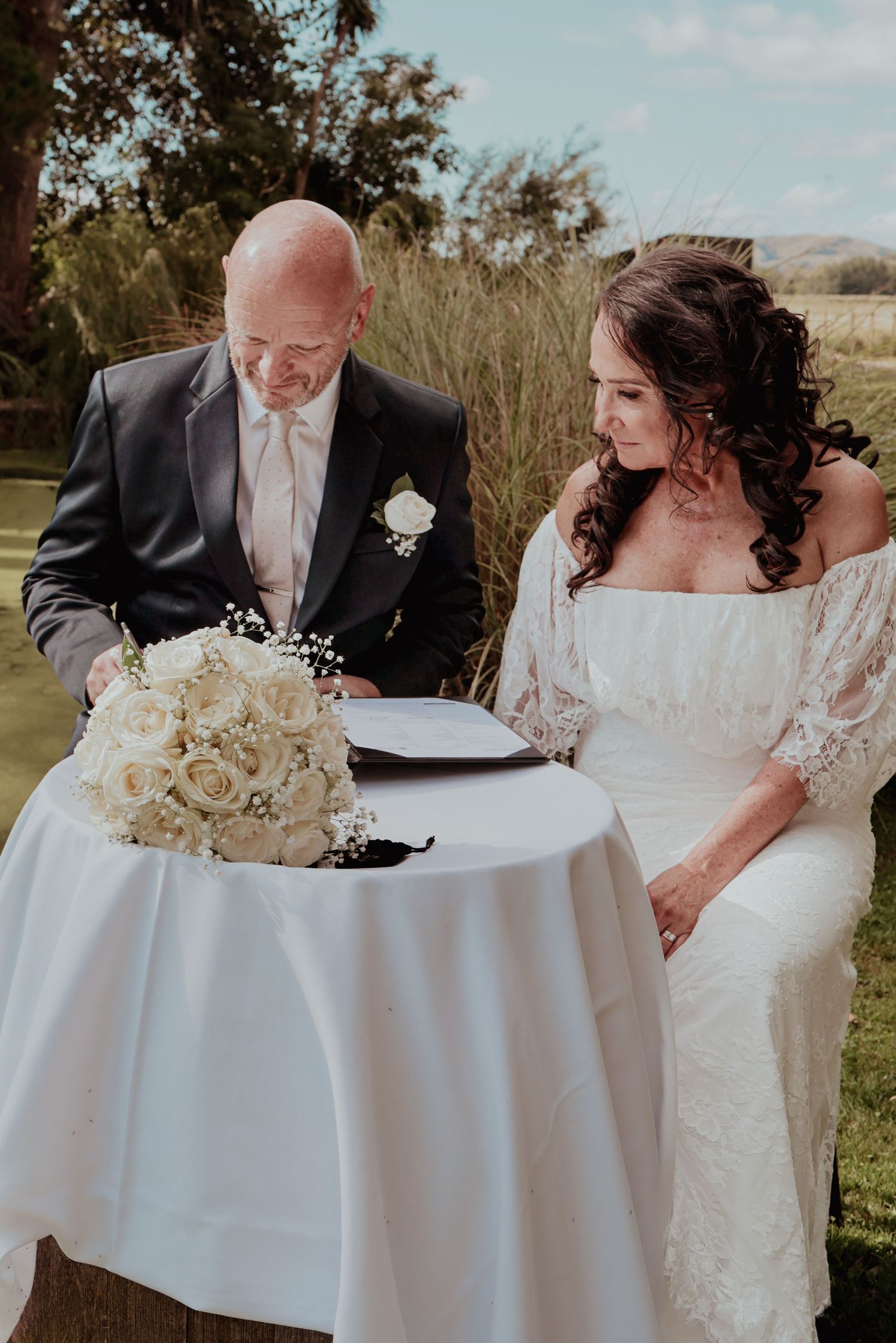 Newlyweds sign wedding register at white draped table with white rose bouquet outdoors.